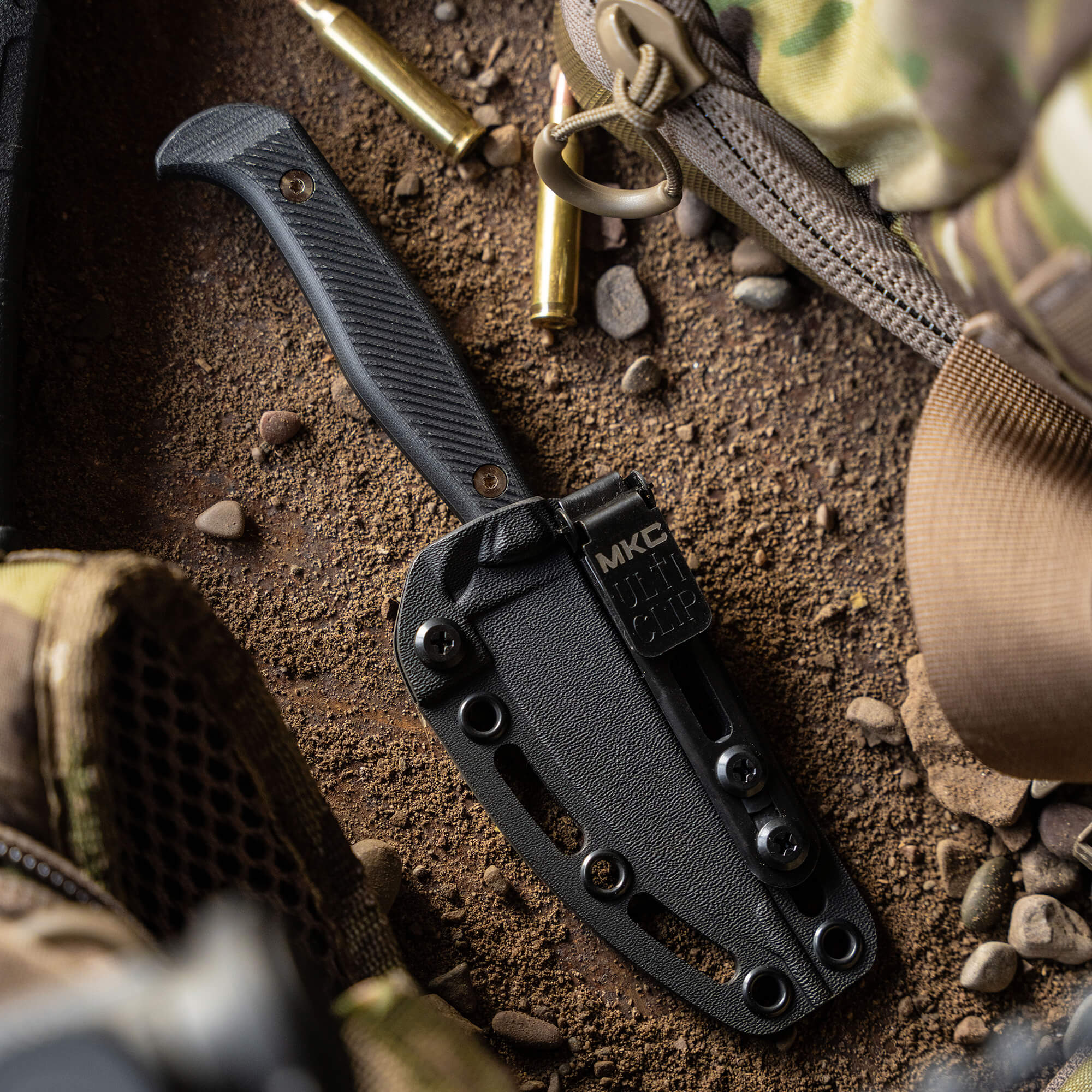 Alt text: Close-up of a black tactical knife, the Battle Goat BLK/BLK, sheathed in a matching black carrier with a textured handle and mounted on a rough terrain with scattered bullets and tactical gear around it.