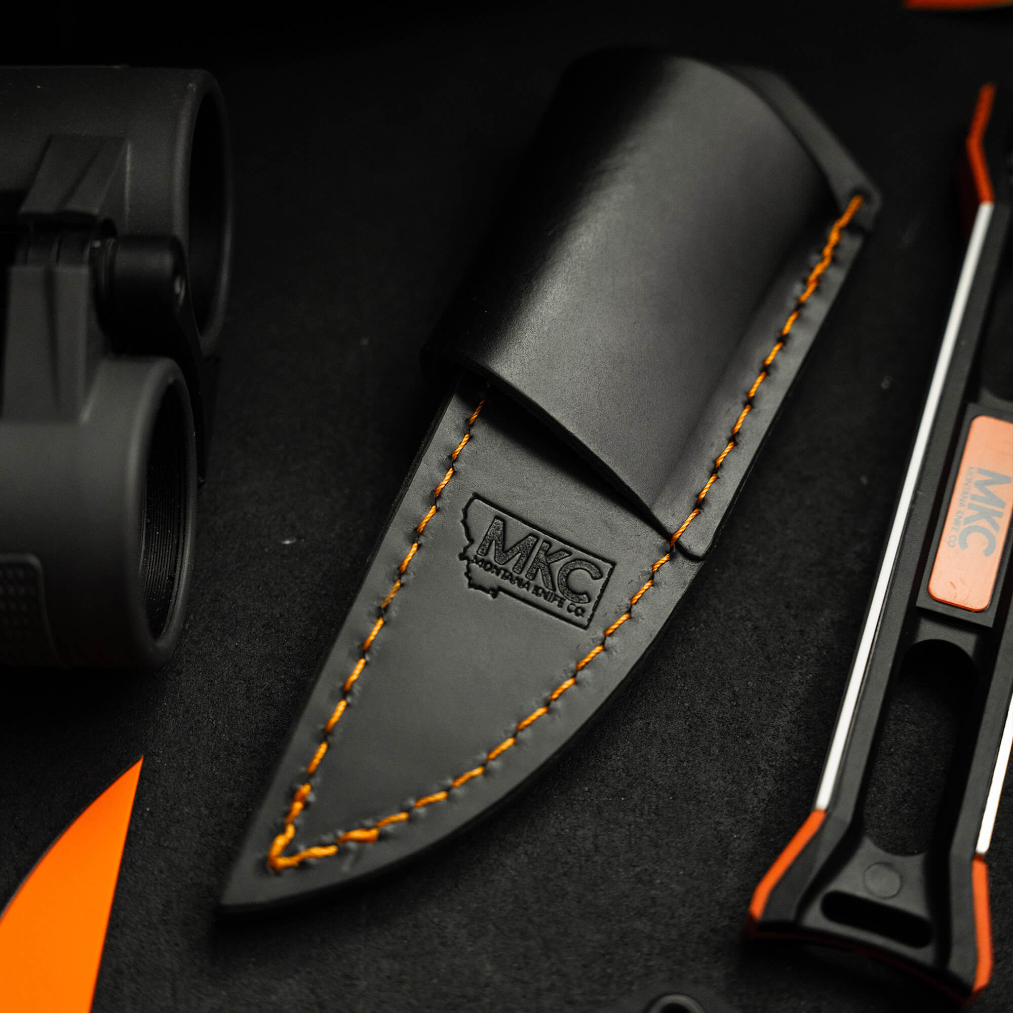 Black leather sheath with orange stitching designed for horizontal belt carry, prominently displaying the MKC logo, featured alongside a tactical knife and outdoor gear on a dark background.