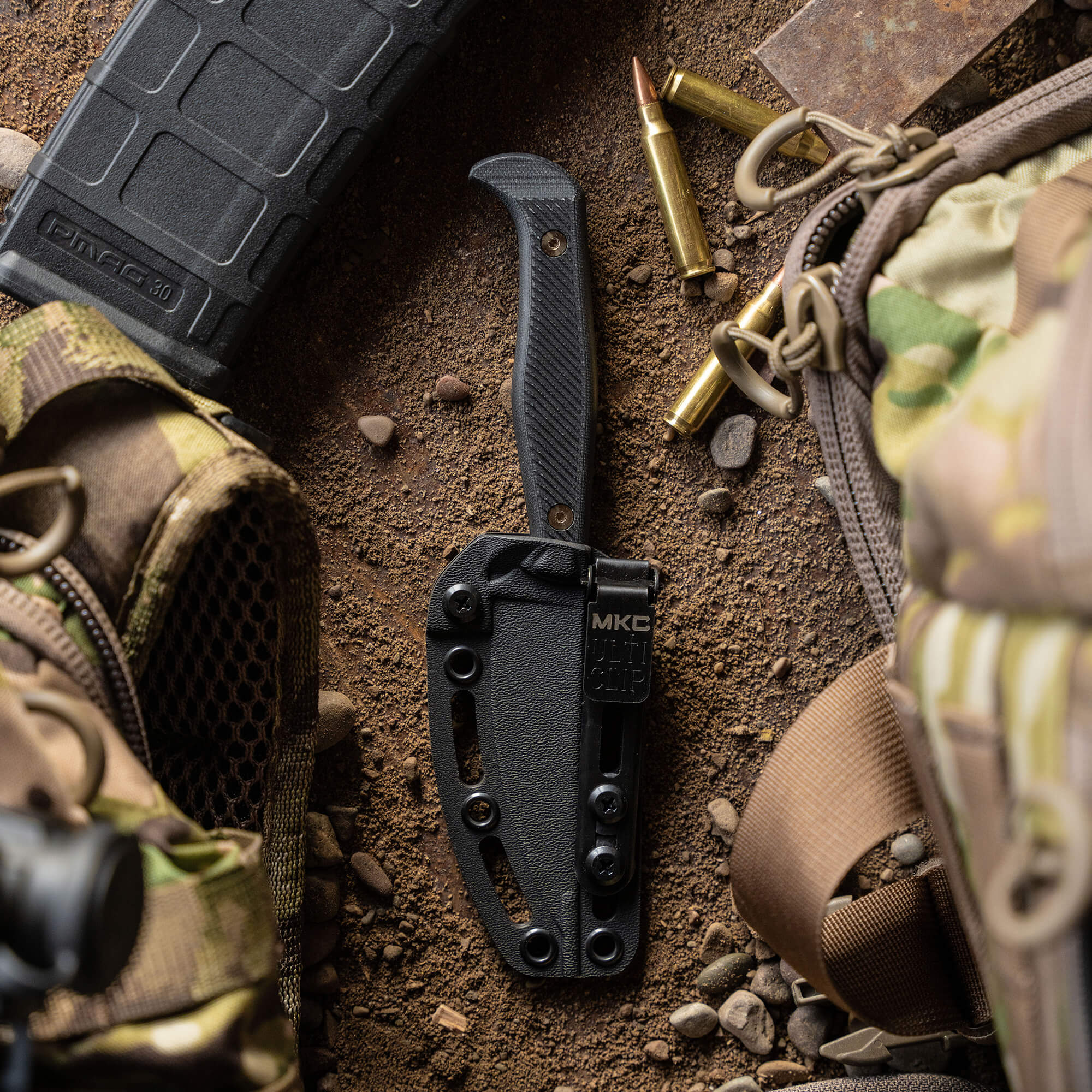 Alt text: "Black tactical knife BATTLE GOAT - BLK/BLK with sheath, surrounded by outdoor equipment including bullets, a tactical backpack, and a firearm magazine on a rough earthy ground."