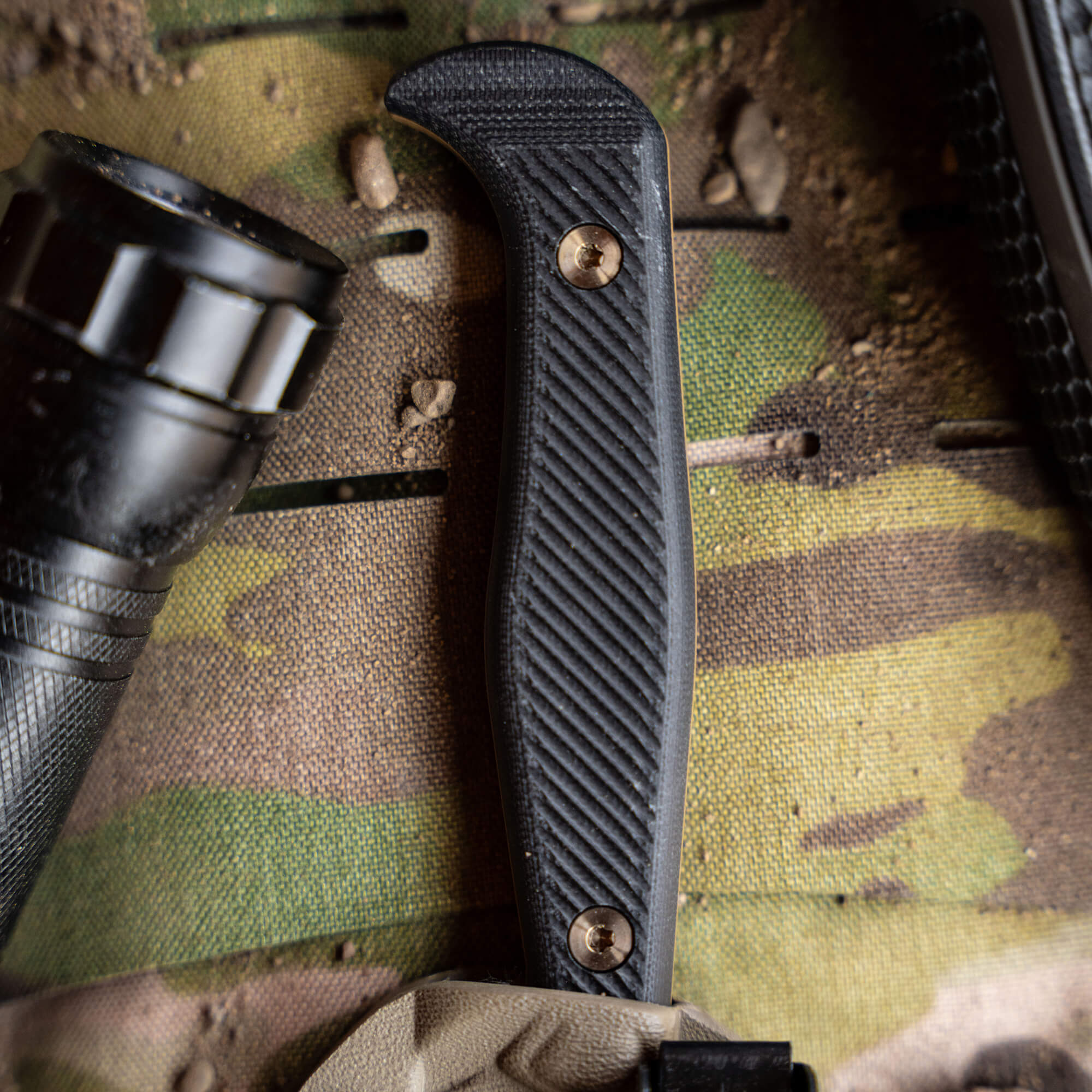 Close-up view of a black tactical knife handle with textured grip, gold screws, set against a backdrop of camouflaged fabric with a flashlight and tactical gear elements around.