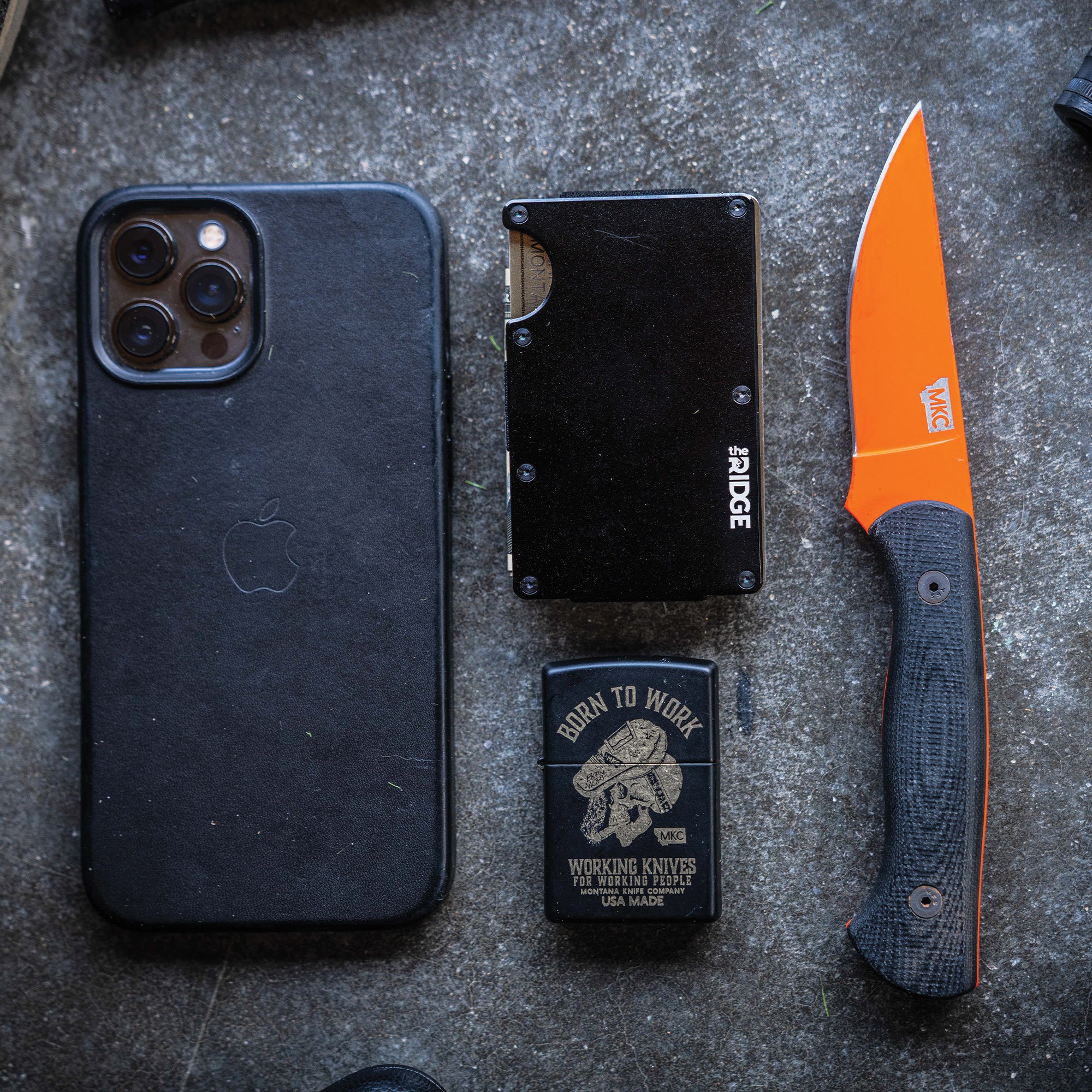 Flat lay of outdoor gear including a black smartphone, a black Ridge wallet with a visible twenty dollar bill, a metallic card featuring MKC branding, and a MKC Blackfoot 2.0 fixed blade knife with a blaze orange blade and black textured