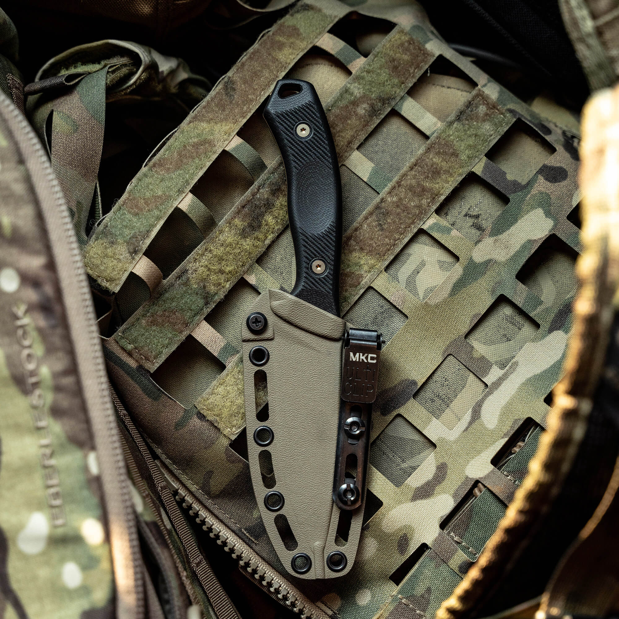 Alt text: "Coyote-colored TF24 Kydex sheath attached to a camouflaged tactical vest holding a black-handled knife, displayed in an outdoor military gear setting."