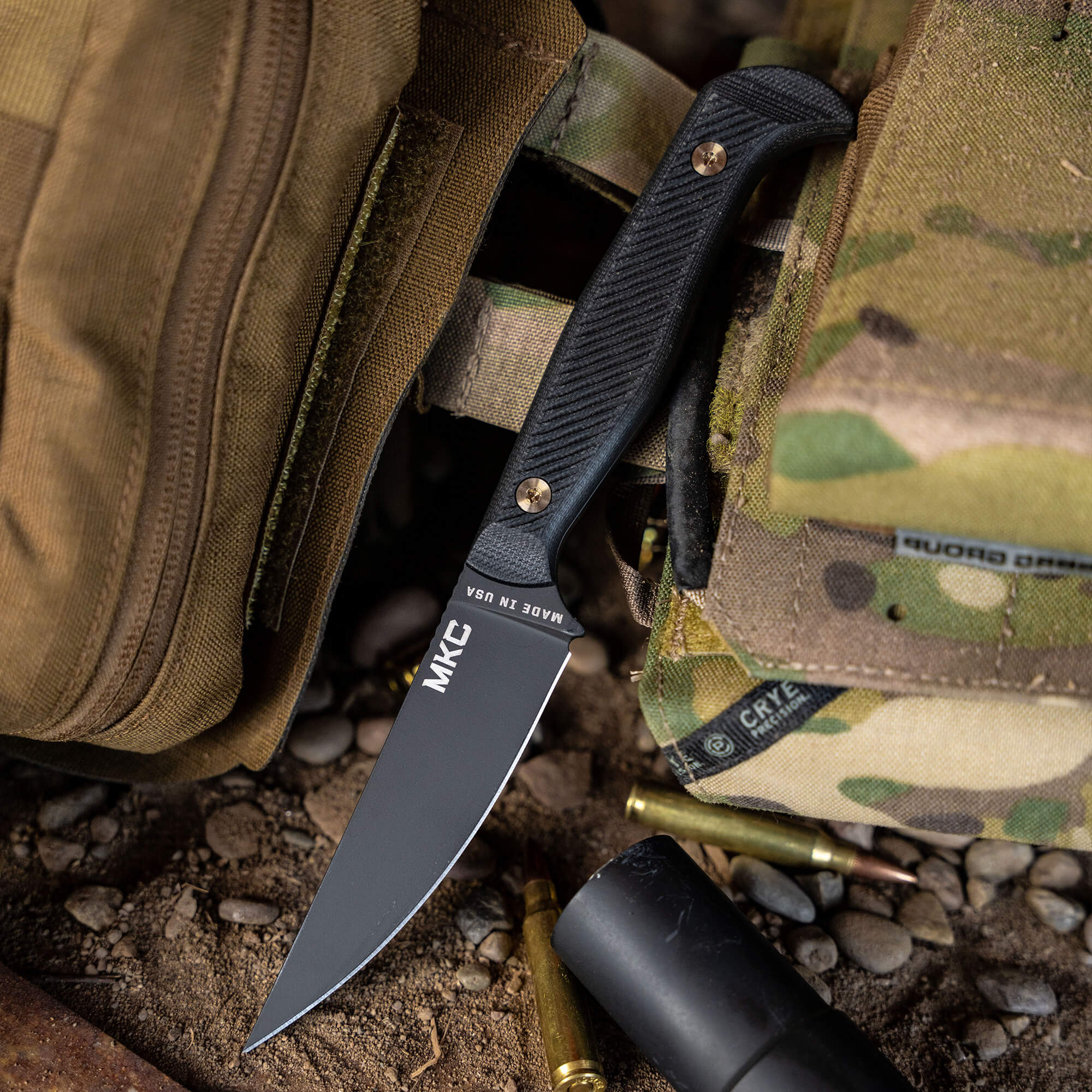Alt Text: "Close-up of the Battle Goat tactical knife in black, featuring a sleek blade and textured handle, partially tucked into camouflage gear with ammo and outdoor equipment nearby, reflecting a rugged and prepared outdoor setting."