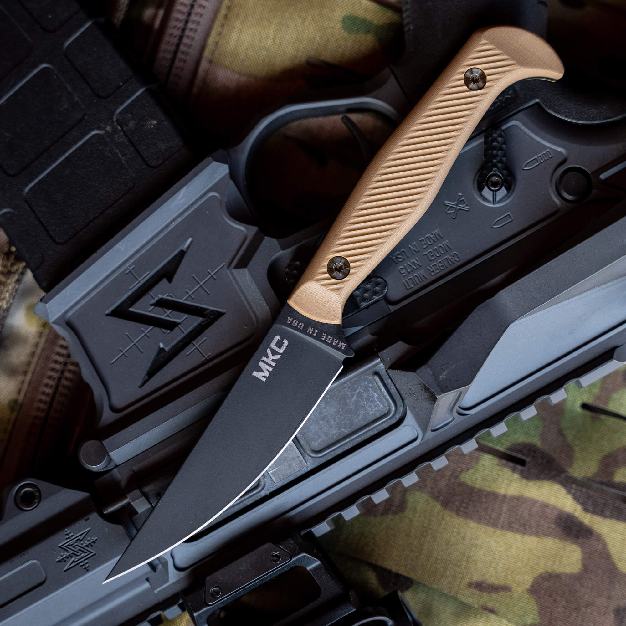 Alt text: "Close-up view of a BATTLE GOAT tactical knife with a smooth black blade labeled MKC and a textured tan handle, resting on a gray and black gun with camo background."