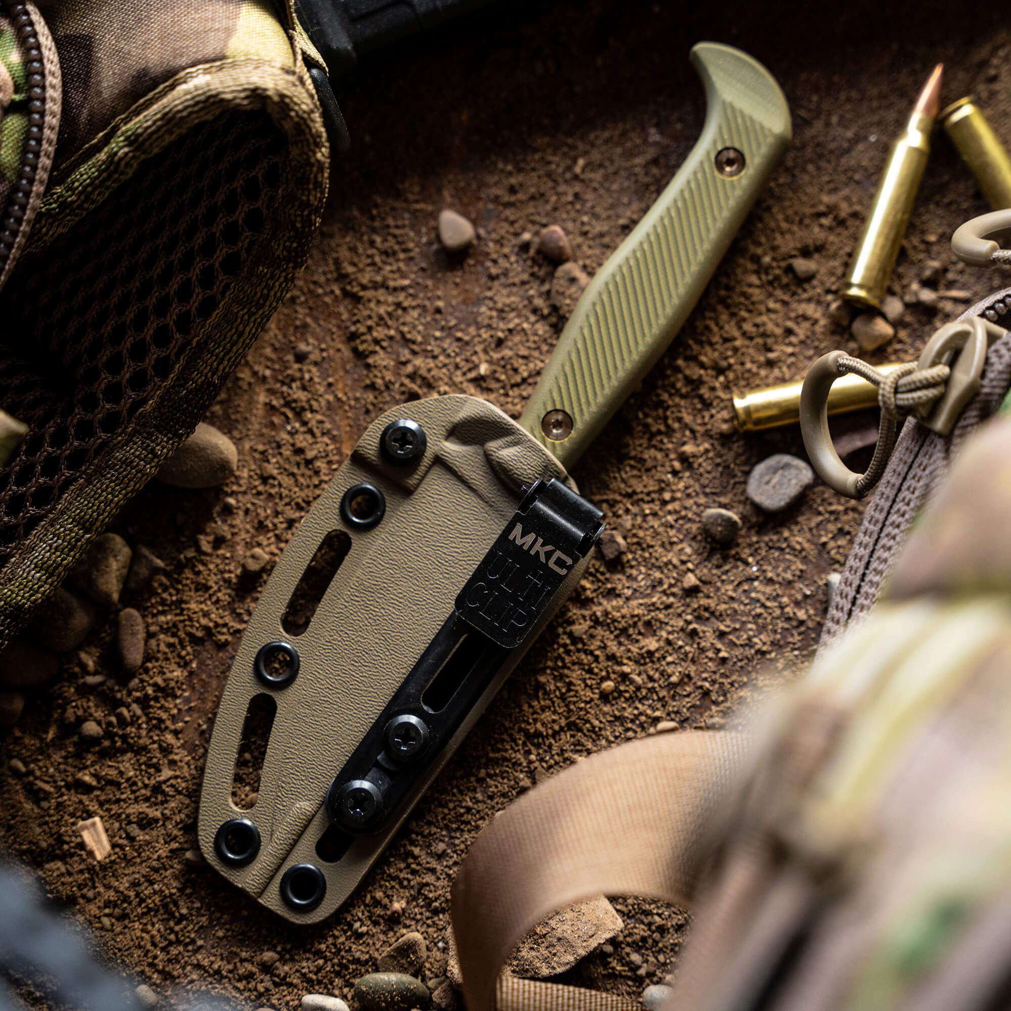 A tactical knife with a coyote tan handle and sheath, displayed on a rugged ground amid military gear and ammunition, highlighting its outdoor utility and durability.
