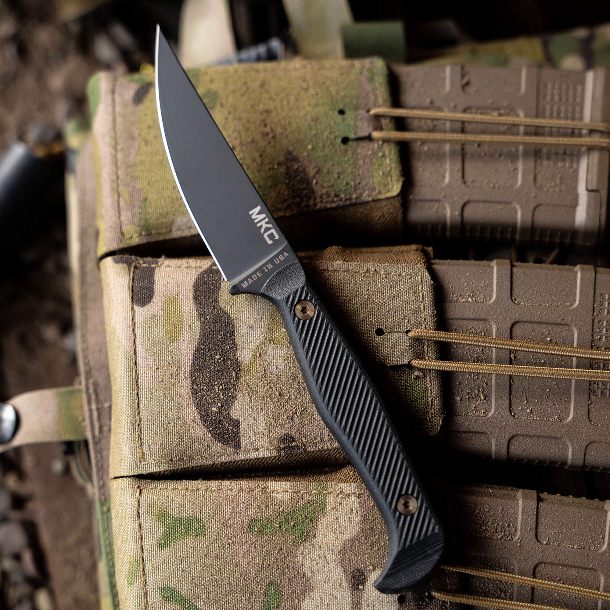 Alt text: A close-up view of a tactical knife, labeled "MKC," placed on a camouflage military tactical vest, emphasizing the knife's black handle and clean blade, with a background of outdoor gear.
