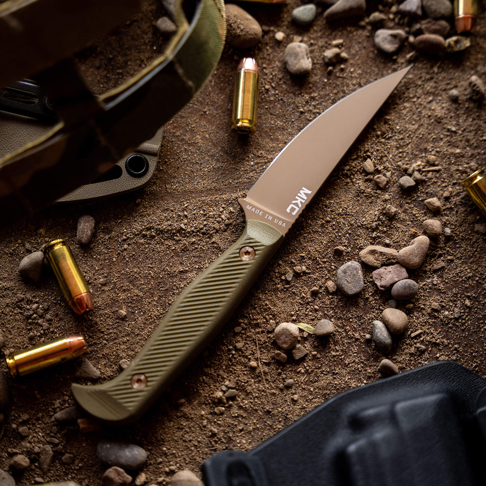 Alt text: "Close-up view of a BATTLE GOAT tactical knife in coyote olive drab color, lying on a gritty surface surrounded by small rocks and spent bullet casings, with partial views of tactical gear and a holster. The blade