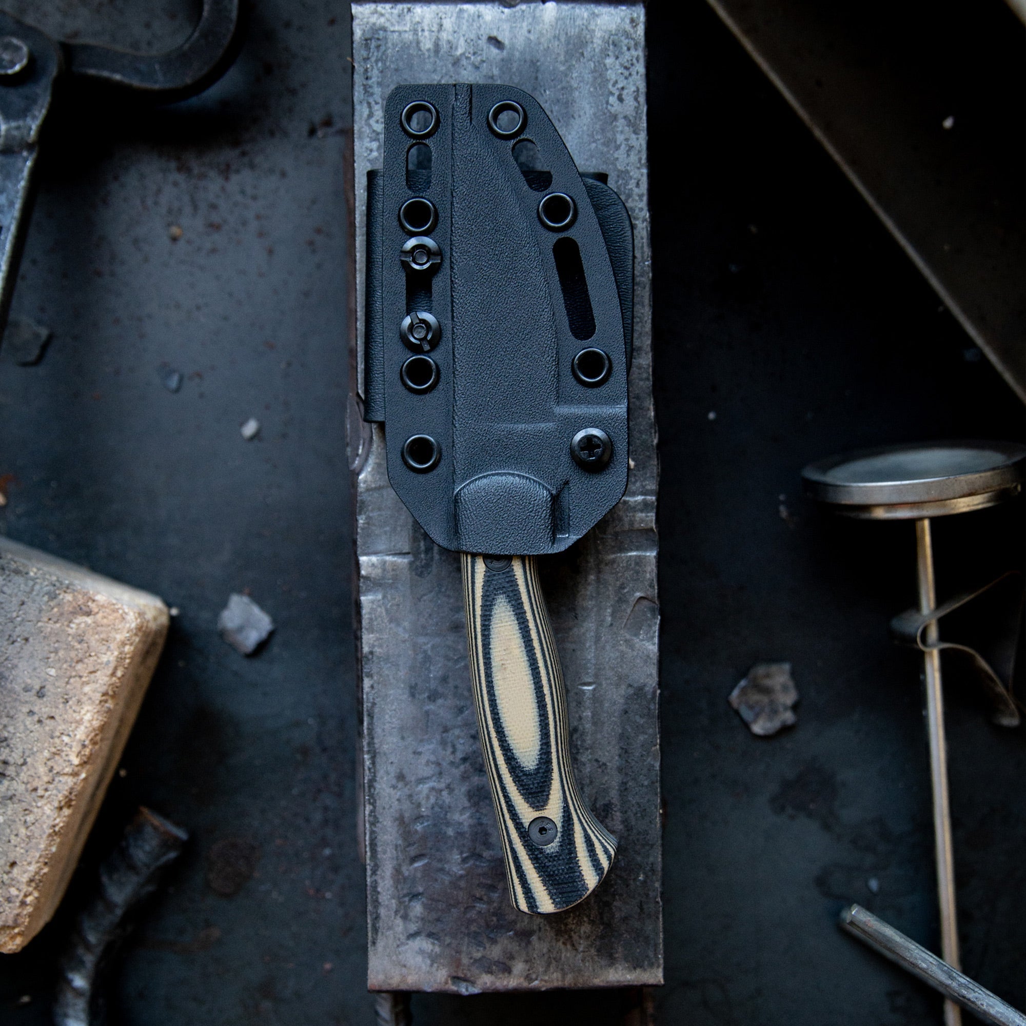 Alt text: Close-up view of the Blackfoot 2.0 knife in a black Kydex sheath, placed on a rustic metal surface in an industrial setting. The sheath features multiple mounting holes and a belt clip.
