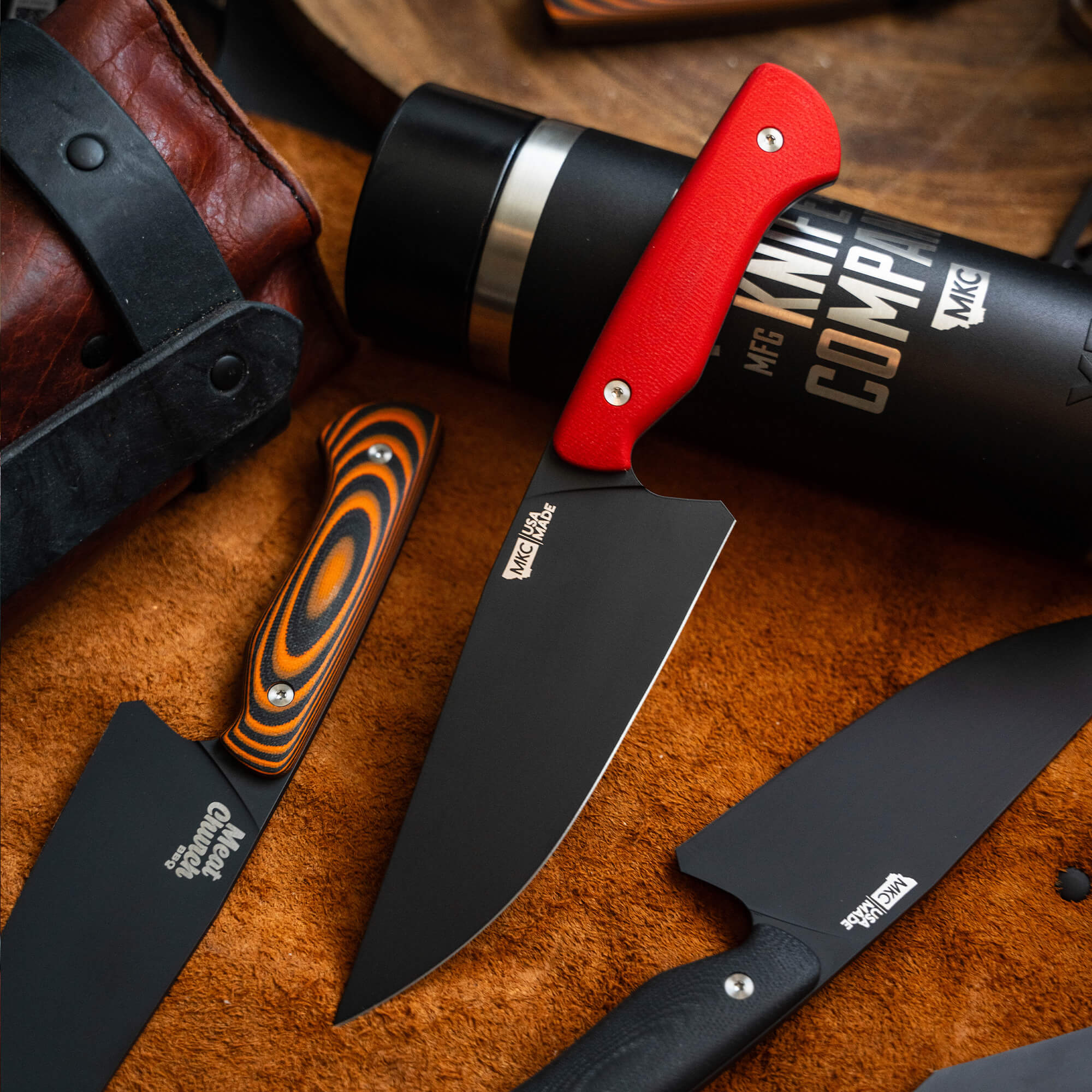 Alt text: "A close-up view of the Meat Church chef knife with a red handle prominently displayed amongst other knives and a black water bottle on a rustic brown leather backdrop."