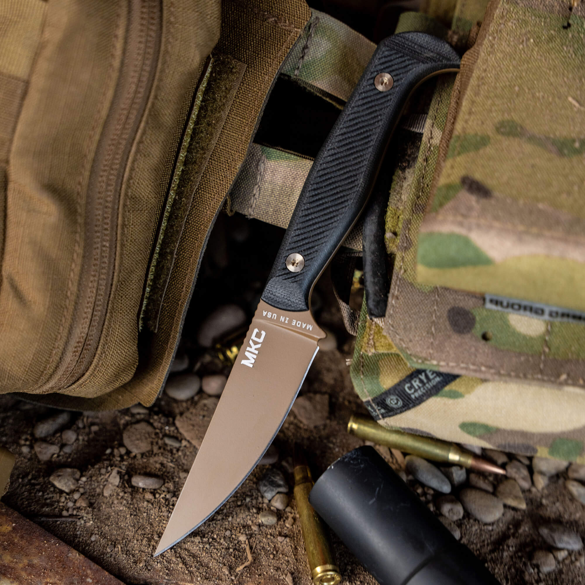Alt text: "Close-up of a MKC Battle Goat folding knife with a black textured handle and a tan blade, partially resting on a rugged khaki bag next to military equipment and ammunition on a gravelly ground."