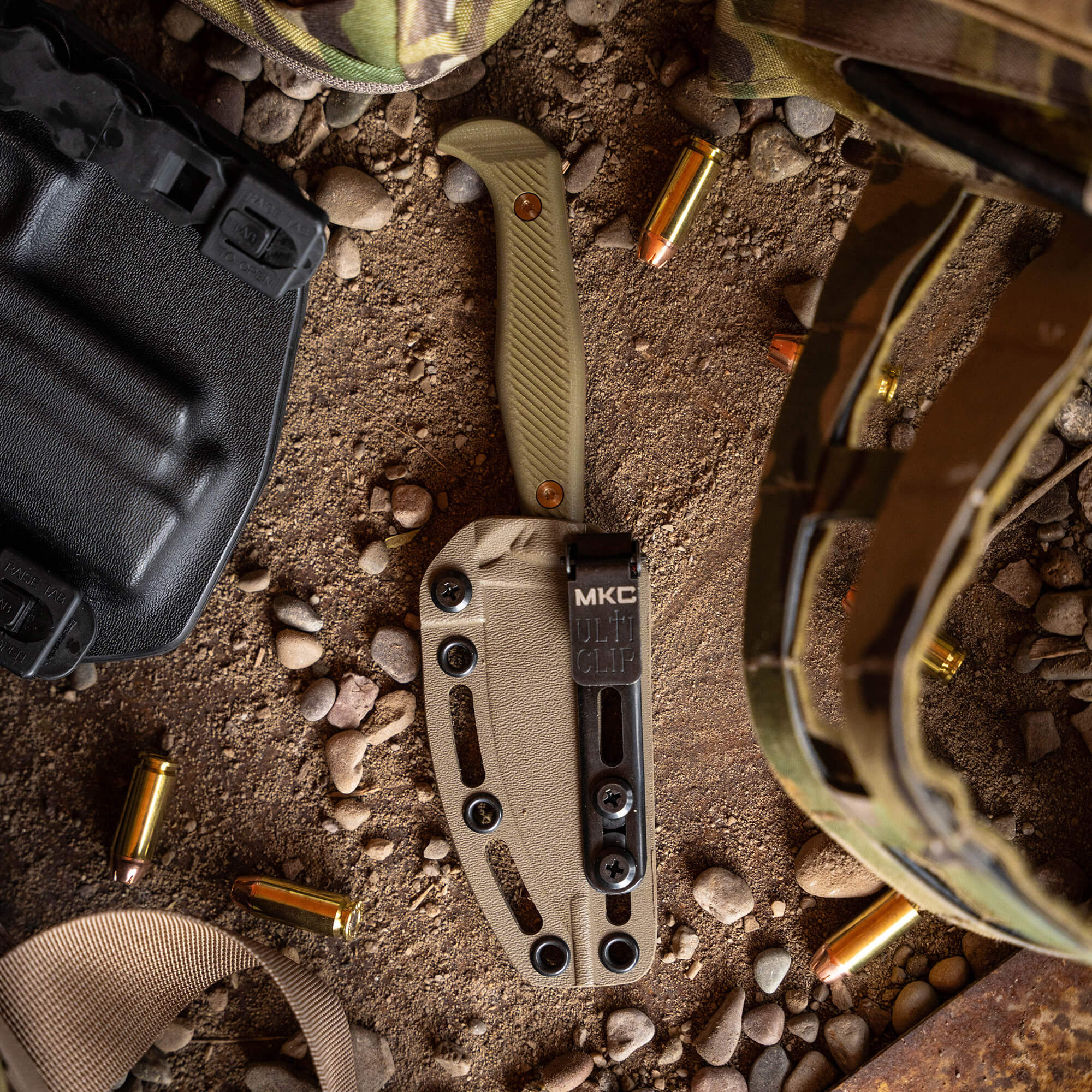 Alt text: "Close-up of a BATTLE GOAT tactical knife in Coyote Olive Drab color, partly unfolded and placed on sandy terrain surrounded by tactical gear items including a helmet, a fabric clip, and scattered bullet casings."
