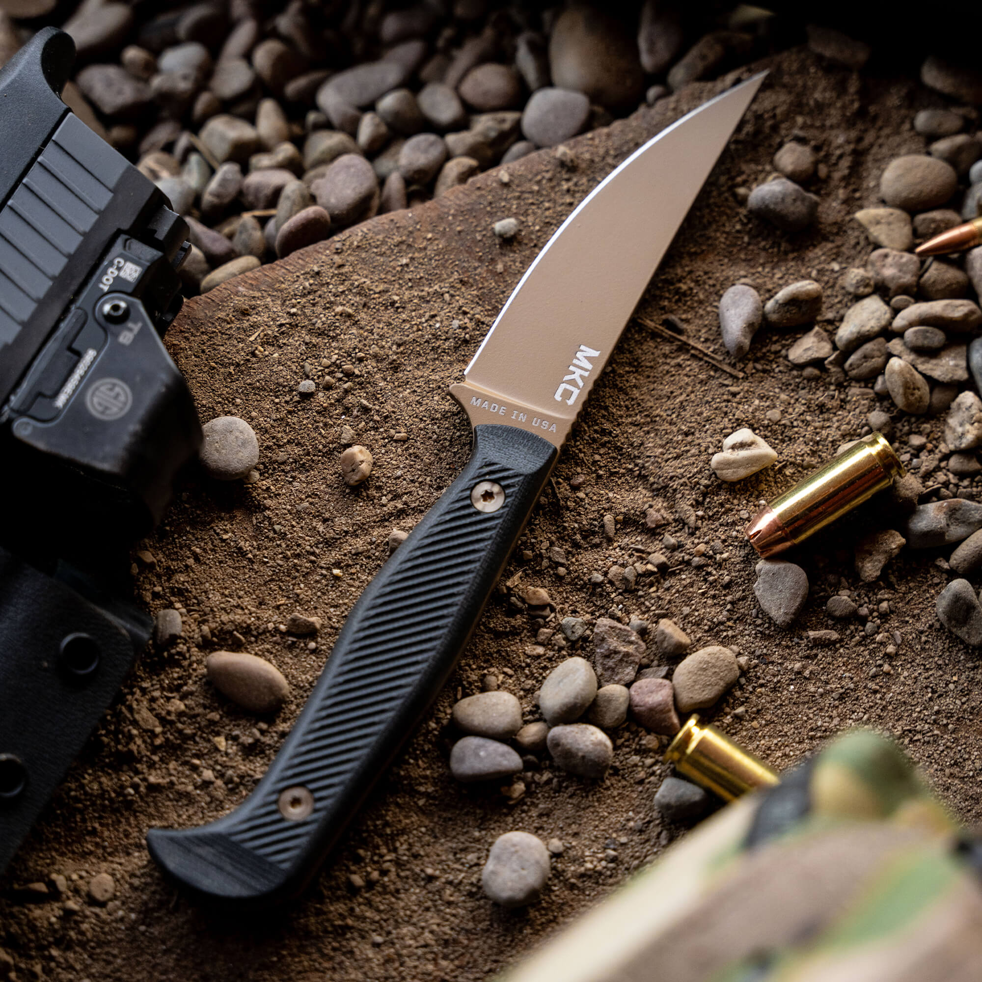 Alt text: "Close-up of a BATTLE GOAT Coyote Black tactical knife with a black textured handle and a silver blade, prominently displayed on a gritty surface surrounded by small stones, bullet casings, and part of a black firearm."