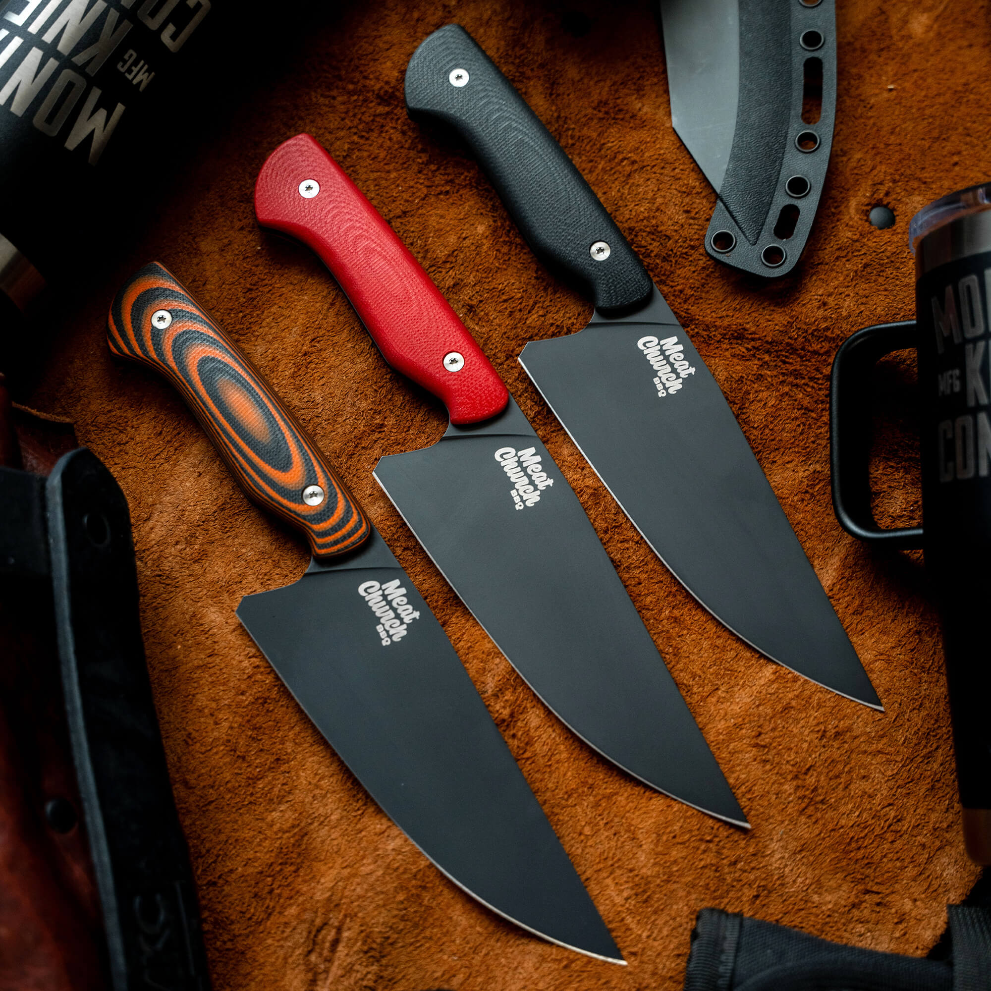 Alt text: "A collection of Meat Church chef knives with various handle colors including red, grey, and multi-colored striped, displayed on a textured brown surface. The knives feature wide, flat blades with the Meat Church logo prominently displayed."