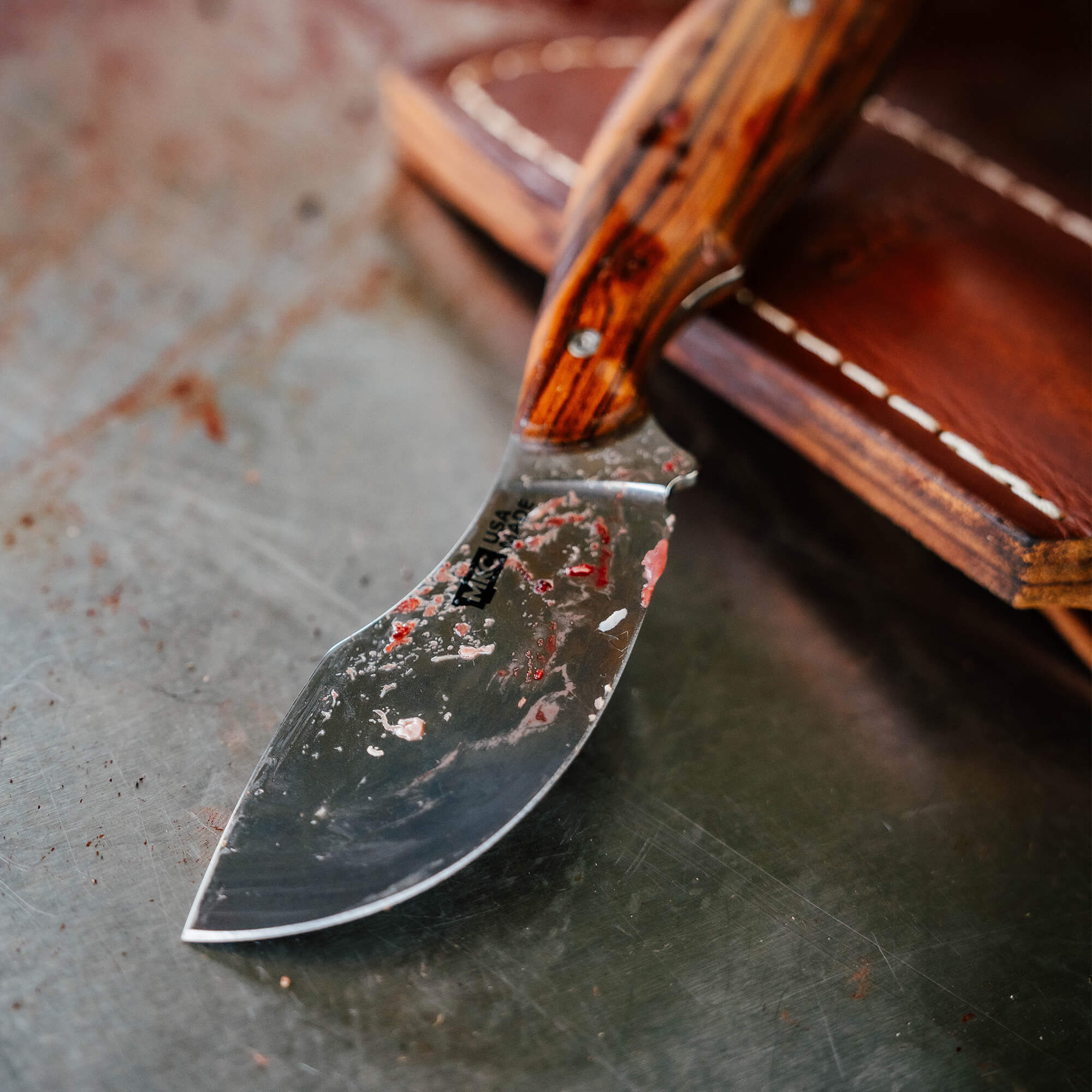 Close-up of the Keep Hammering Traditions Packout Skinner knife with a stained blade and wooden handle, resting on leather.