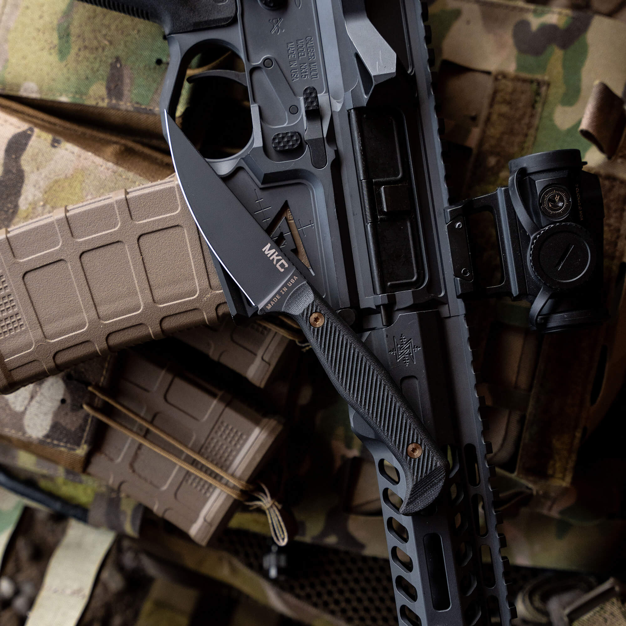 Close-up image of the BATTLE GOAT tactical knife in black, mounted on an assault rifle with detailed markings and accessories, set against a background of military gear and camouflage patterns.