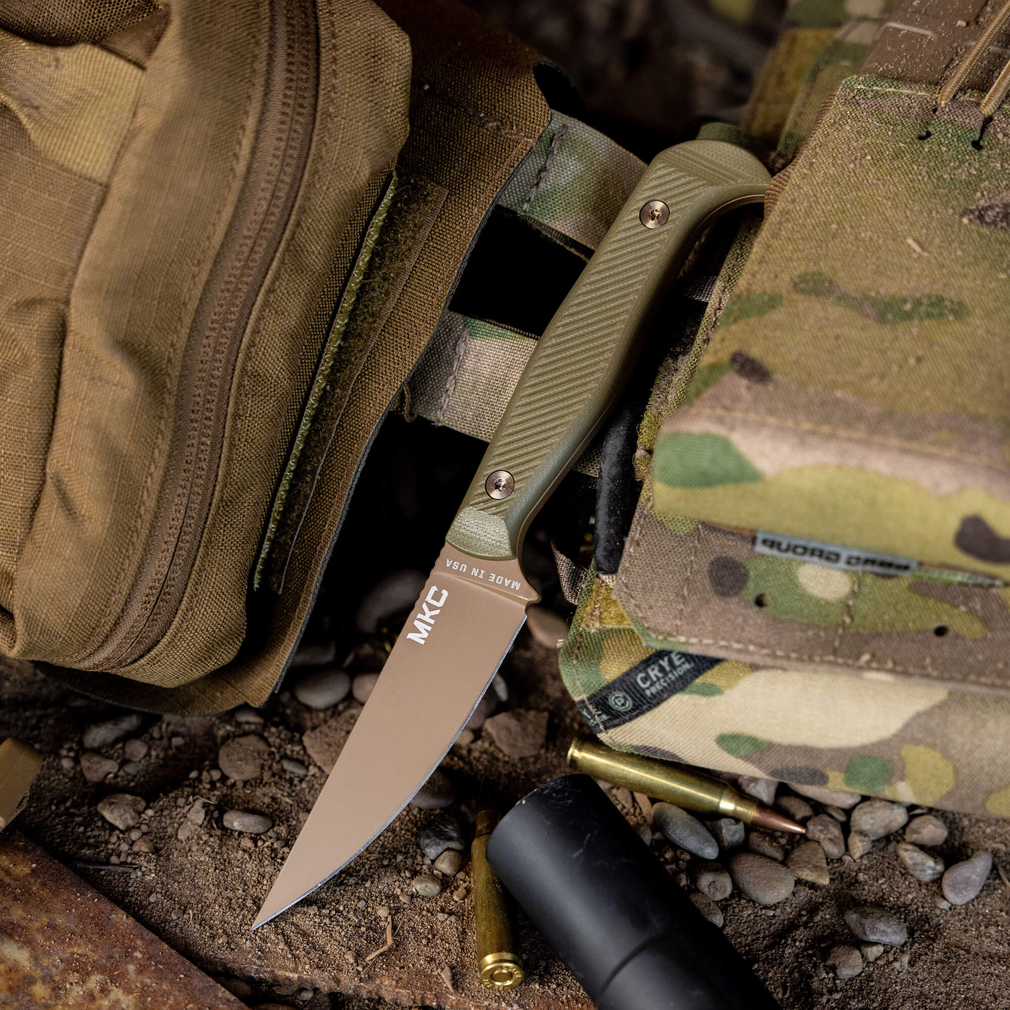 Alt text: Olive green BATTLE GOAT tactical knife with a coyote brown blade, partially inserted in a camouflaged military bag among outdoor gear like ammunition and a black cylindrical object, set against a gritty, stony background.