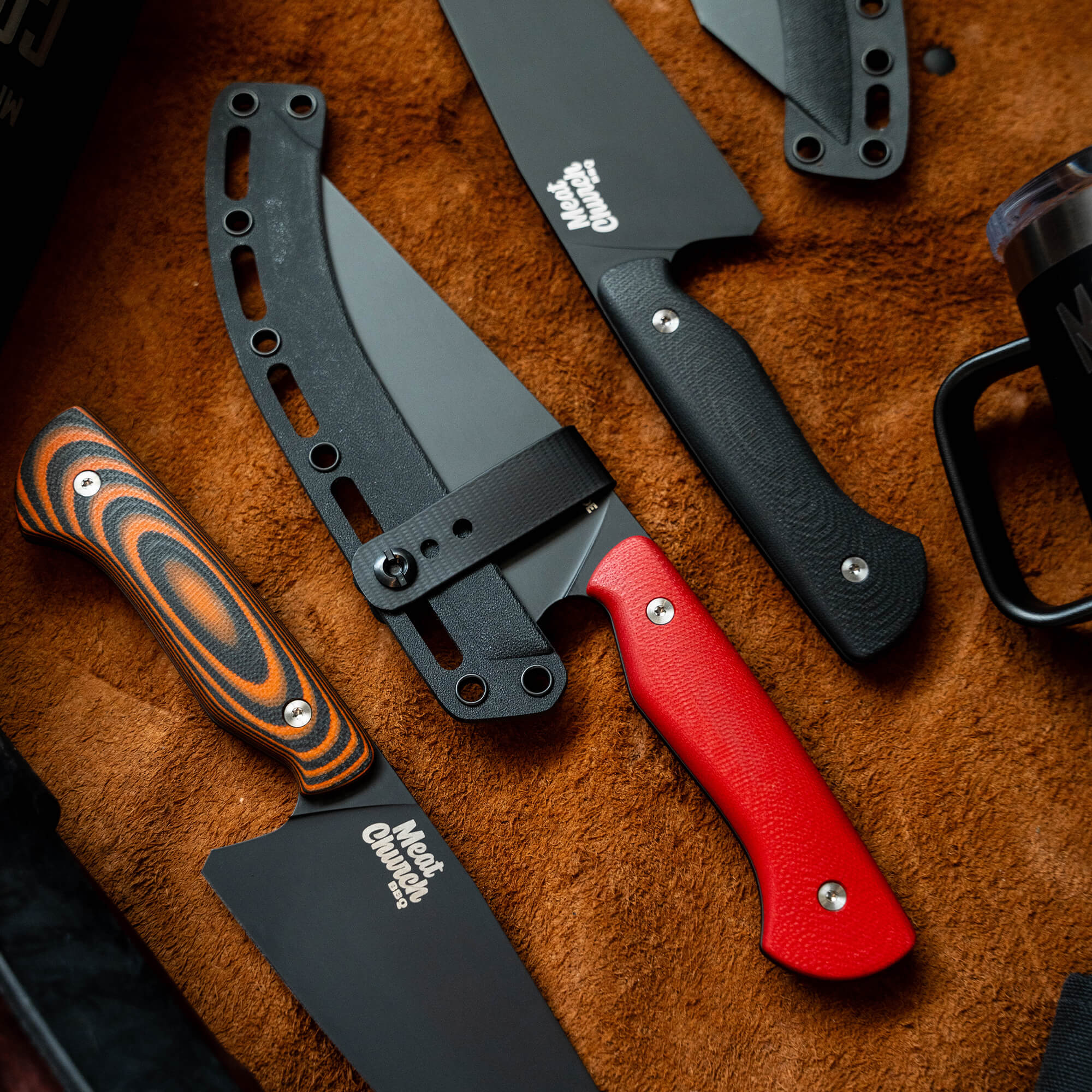 Alt text: "Assorted Meat Church chef knives displayed on a textured brown surface, featuring a prominent knife with a striking red handle and black blade, alongside others with black and multi-colored handles and sheaths."