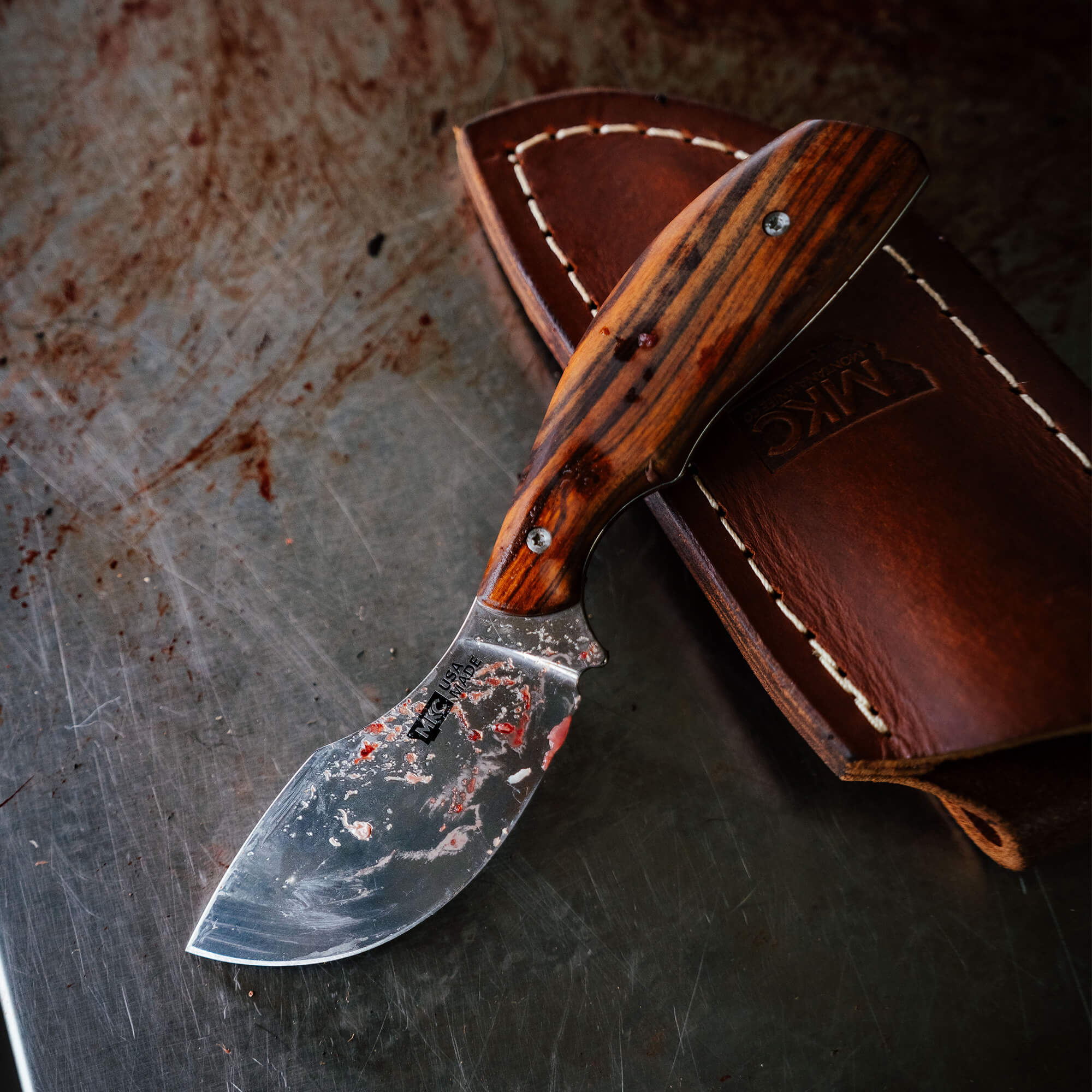 Montana Knife Company's Keep Hammering Traditions Packout Skinner with a wooden handle and leather sheath on a stainless steel surface.