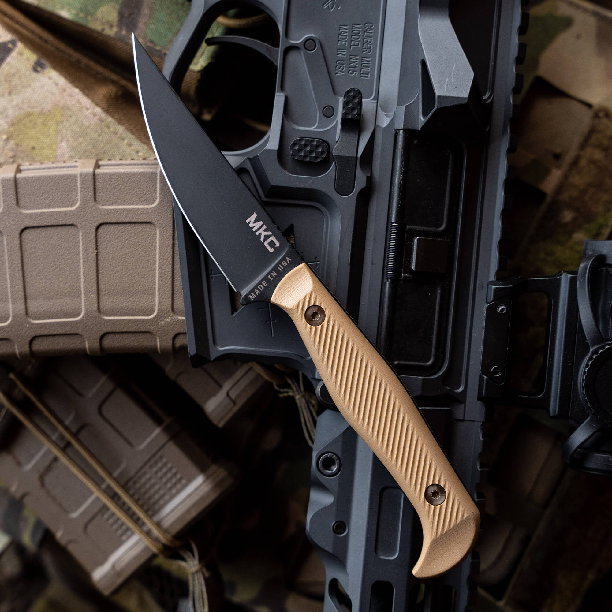 Alt text: "Close-up view of a MKC Battle Goat tactical knife with a black blade and coyote tan handle, placed on a tactical gear setup with elements like firearm magazines and a camouflage background."