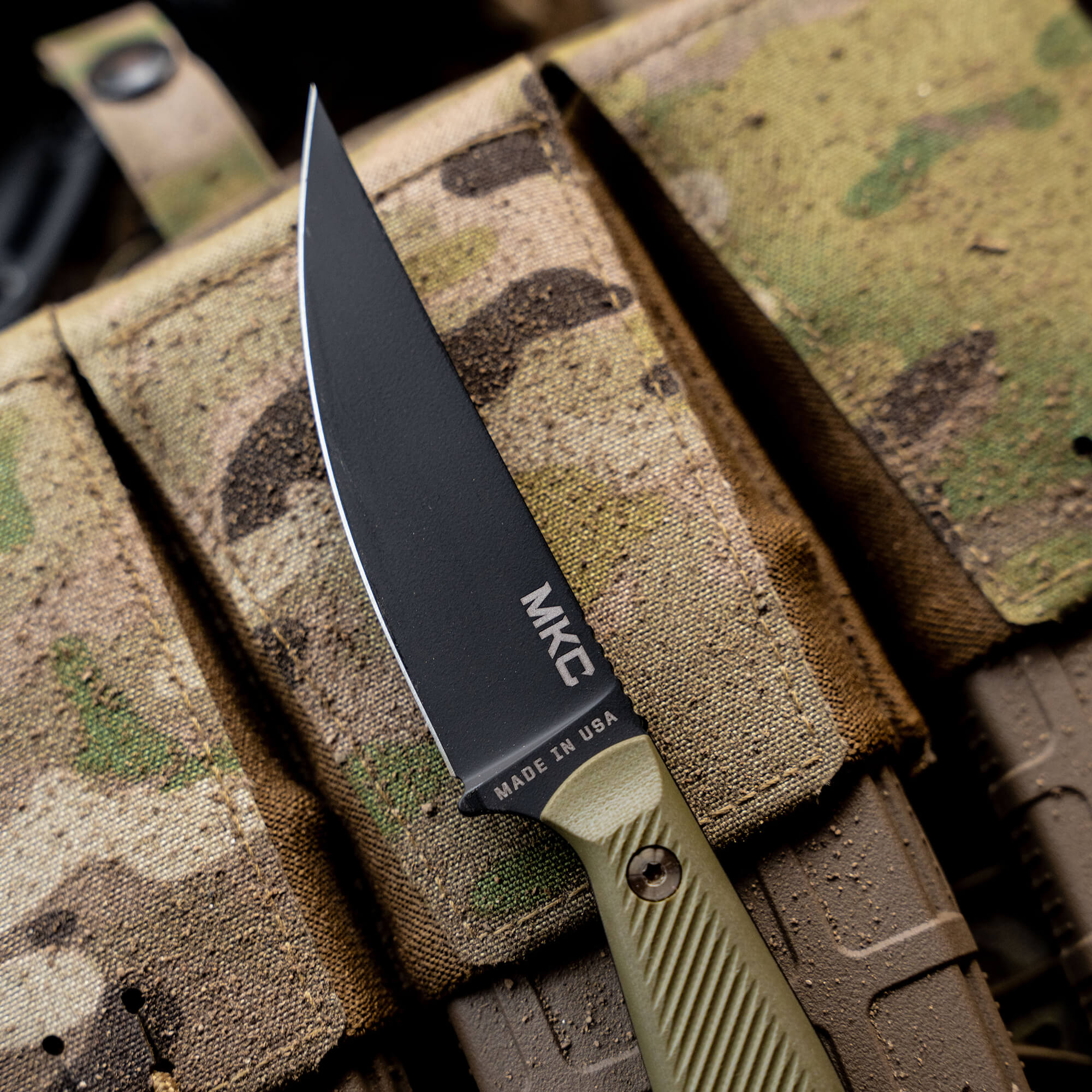 Close-up image of a tactical knife "BATTLE GOAT - BLK/OD" with a matte black blade and olive drab handle, marked with the logo "MKC" and "Made in USA", displayed atop a camouflage fabric background.