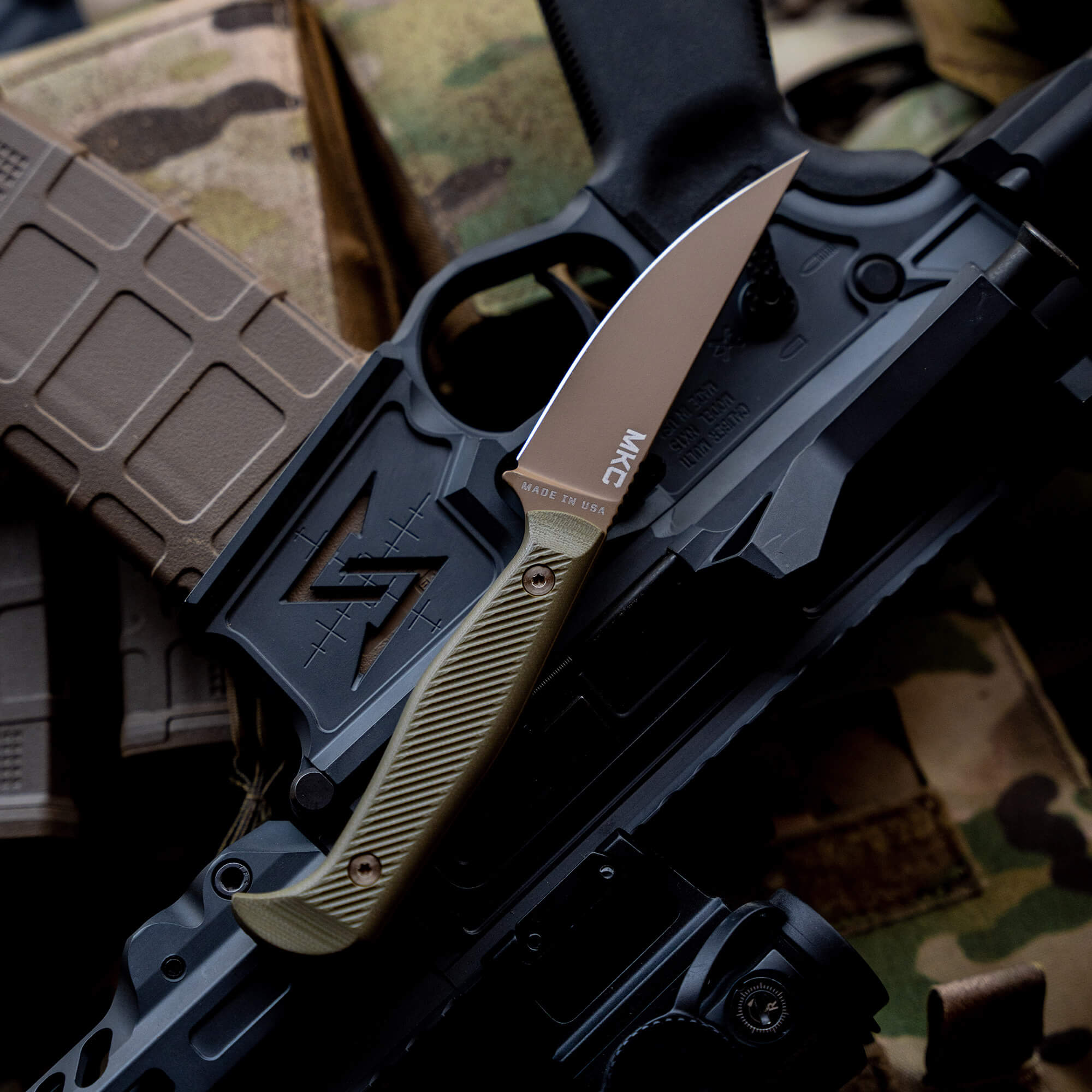 Alt text: "Close-up of a Battle Goat tactical knife in coyote olive drab color, resting on an AR-15 rifle with camouflaged equipment in the background, showcasing its detailed design and robust build."