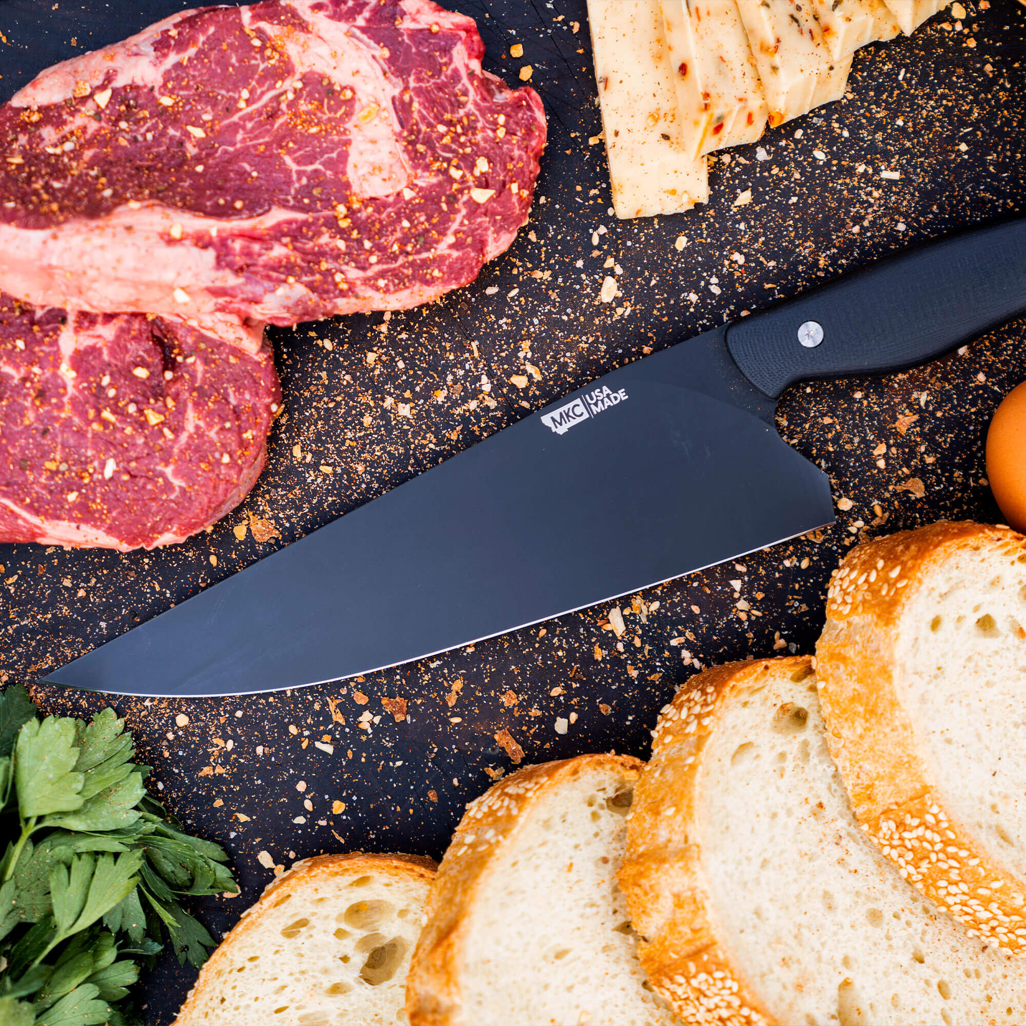 Alt text: A Bighorn Chef black knife with a sleek blade displayed on a textured surface alongside a raw, seasoned steak, sliced cheese, an egg, fresh parsley, and slices of seeded bread.