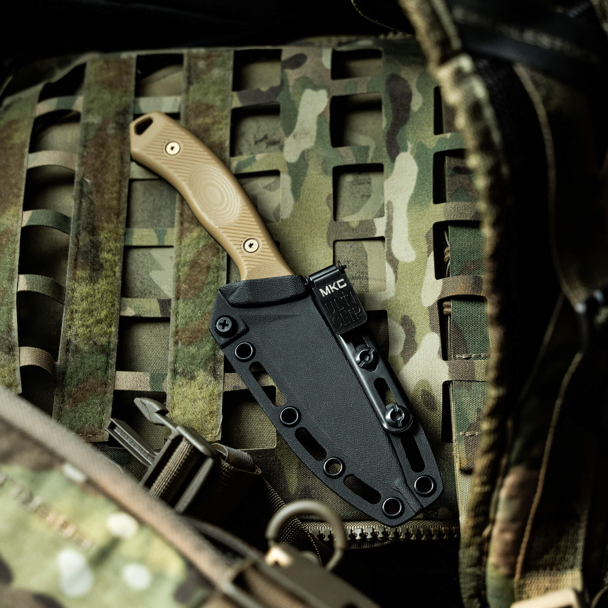 Alt text: "TF24 Kydex sheath in black, featuring a secure and sturdy design, displayed with a tan handled knife, partially sheathed. The sheath is set against a camouflage background, emphasizing its tactical utility."