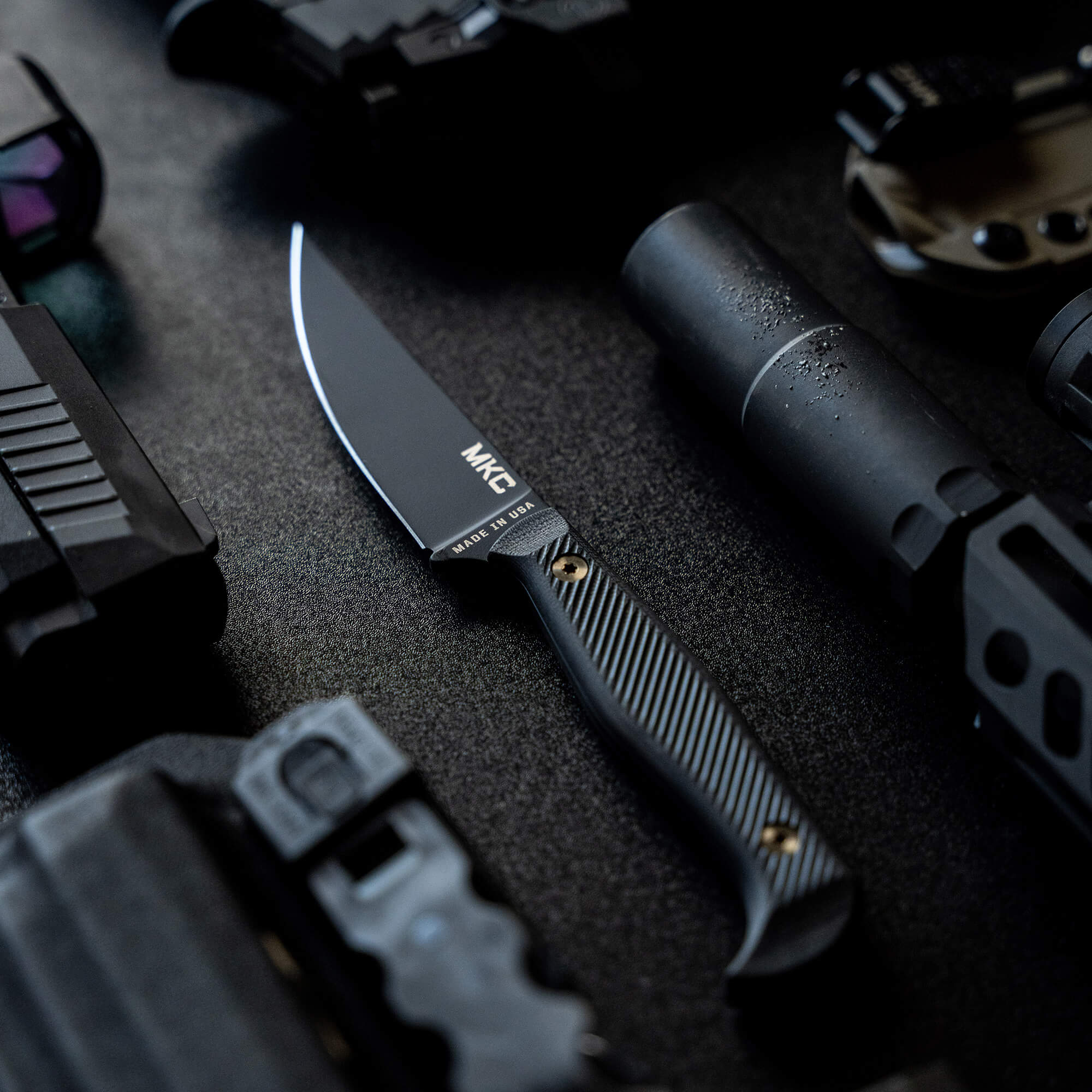 Black MKC Battle Goat tactical knife with textured handle, prominently displayed among various tactical gear items on a dark surface.
