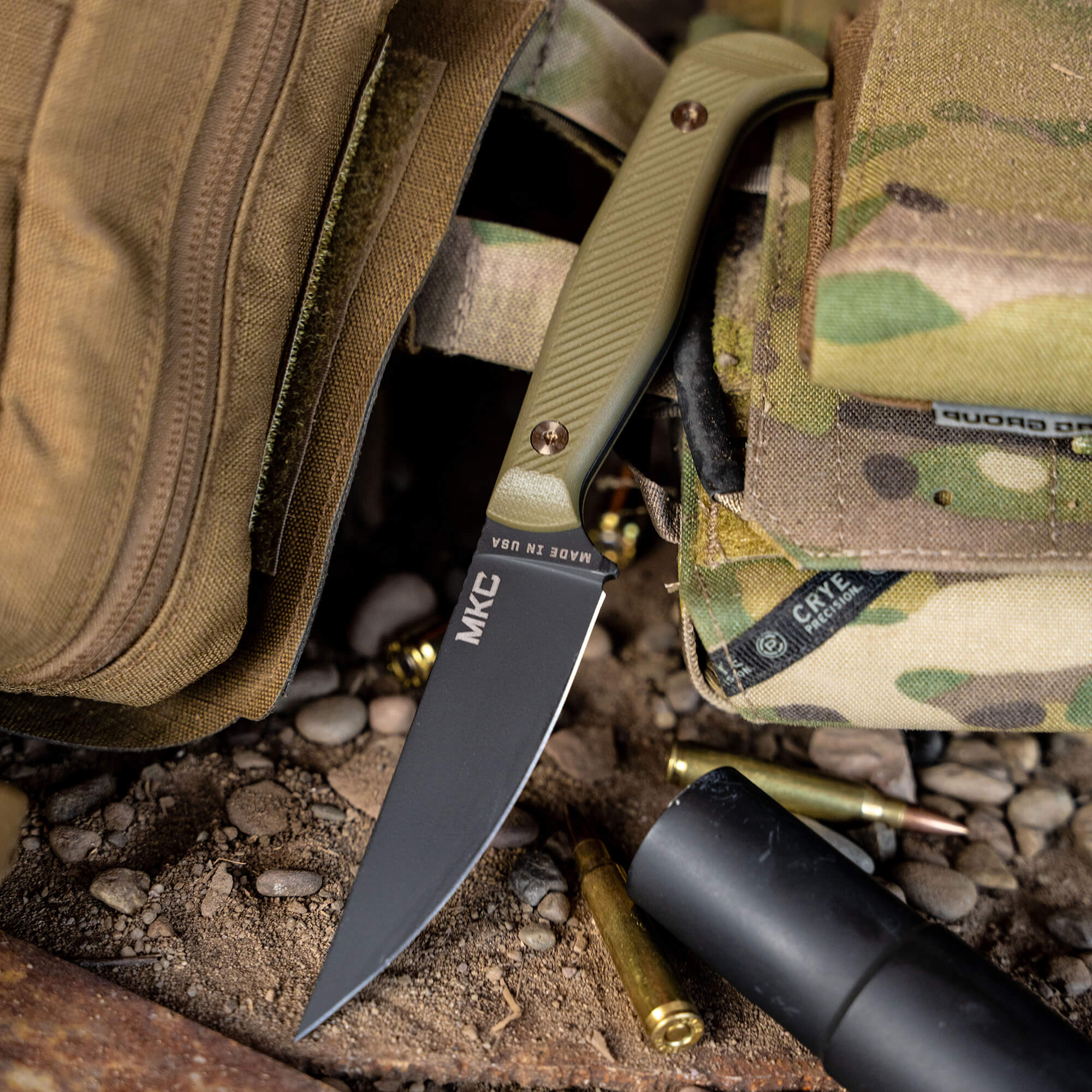 Alt Text: "Close-up of a tactical knife, the BATTLE GOAT - BLK/OD, with a black blade and olive drab handle partially tucked into a camouflaged fabric bag beside spent bullet casings and a flashlight on a