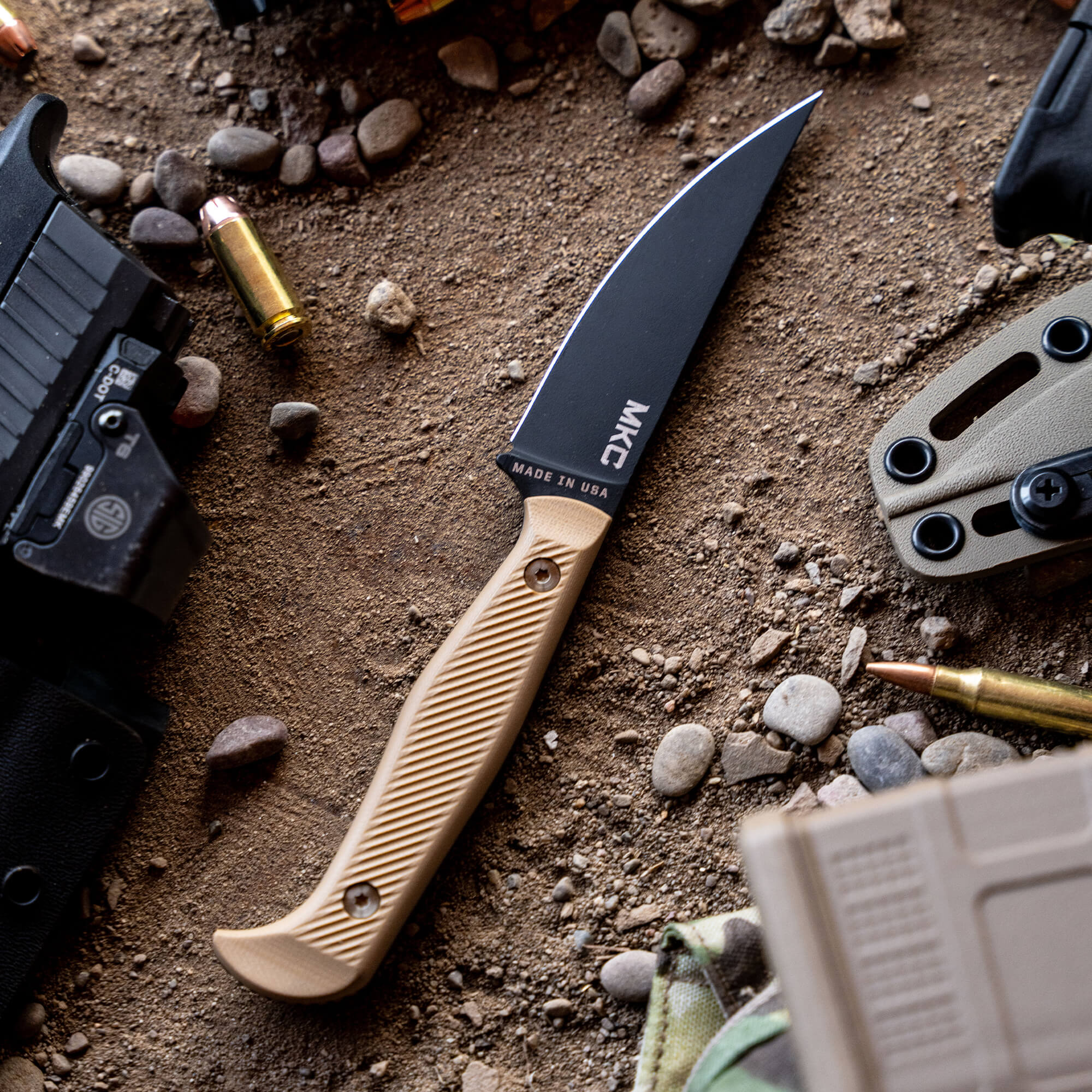 Alt text: "Close-up view of a BATTLE GOAT tactical knife with a black blade and coyote tan handle, marked 'MADE IN USA', surrounded by outdoor gear including a handgun, bullet casings, and tactical equipment on a sandy