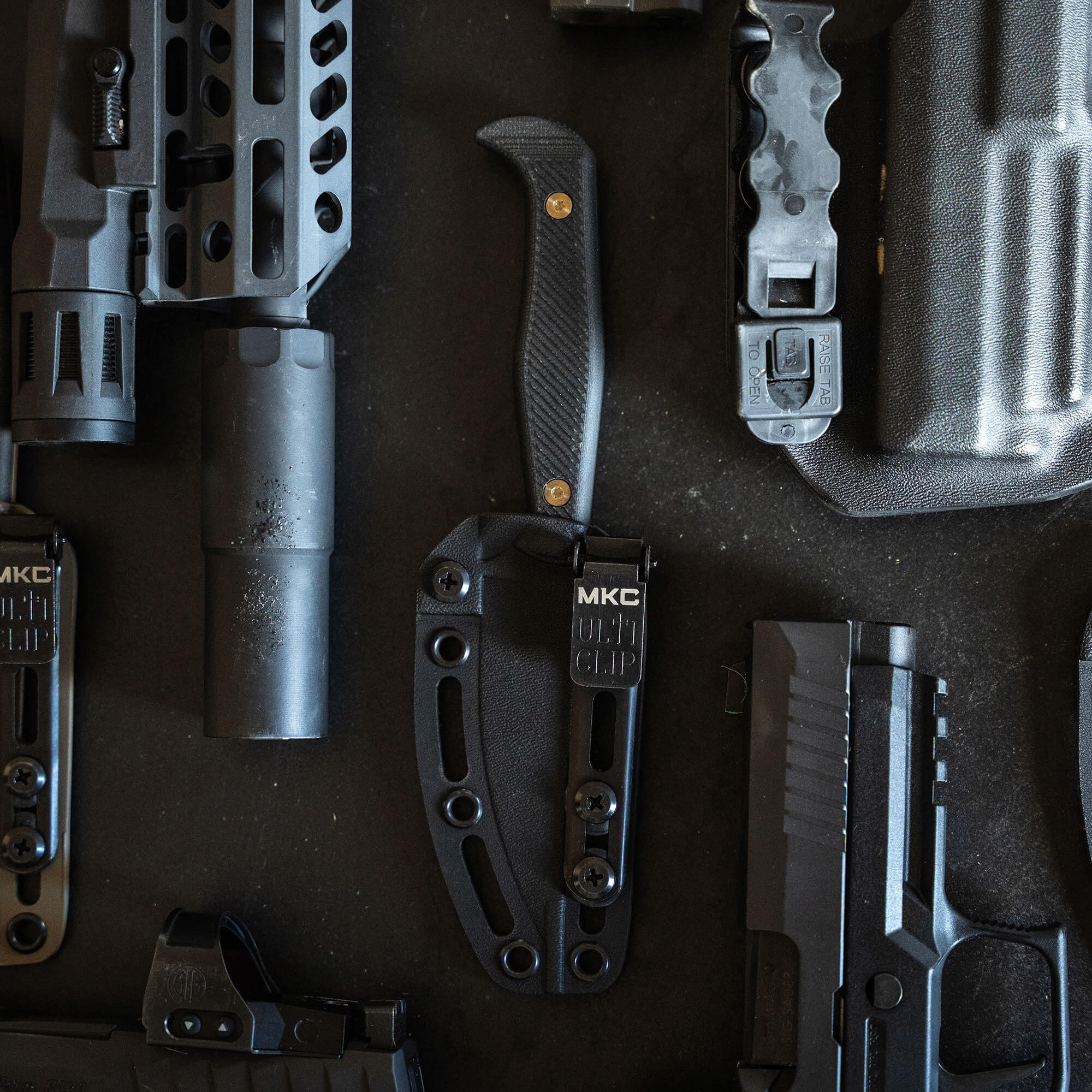 Alt text: "Close-up view of tactical gear including a black combat knife in a sheath, parts of firearms, and various accessories arranged on a dark surface, all in black coloration."