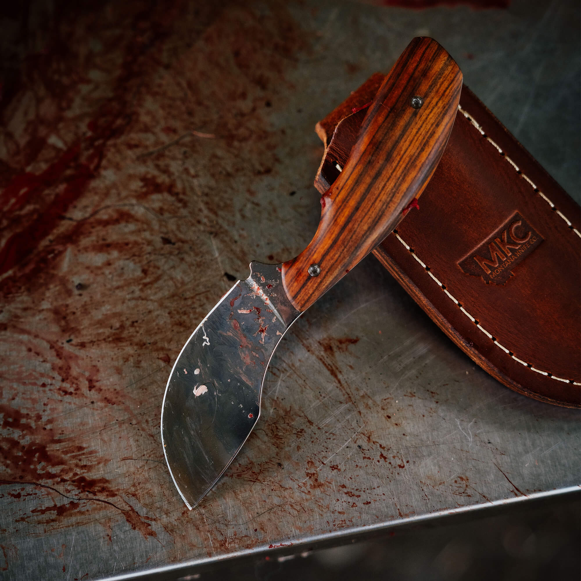 Montana Knife Company's Keep Hammering Traditions Packout Skinner features a sleek blade and a wooden handle, displayed with a leather sheath.