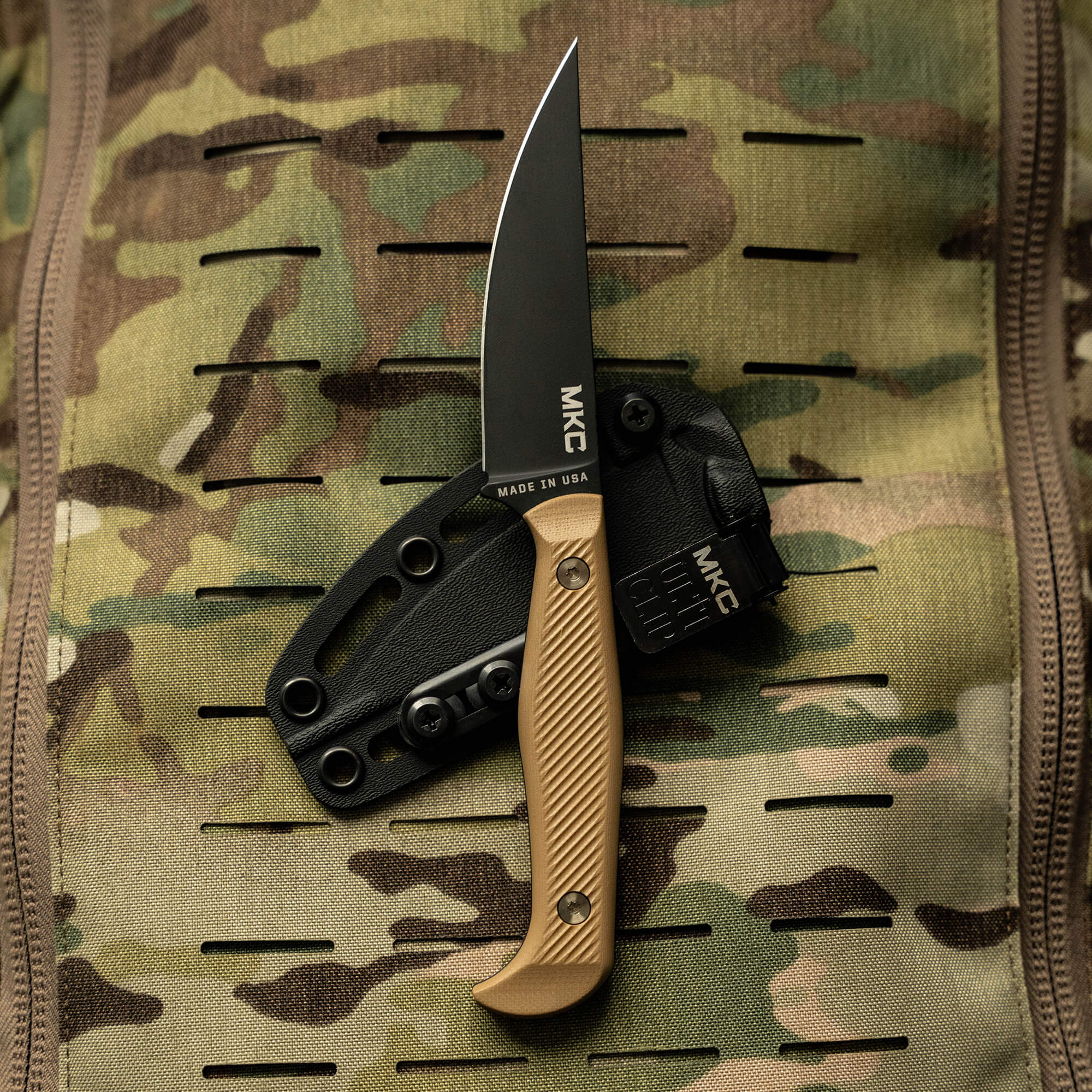 Alt text: "Close-up of the BATTLE GOAT tactical knife with a black blade and coyote brown handle, displayed on a camouflage background. The knife is attached to a black kydex sheath with multiple mounting holes, labeled as made