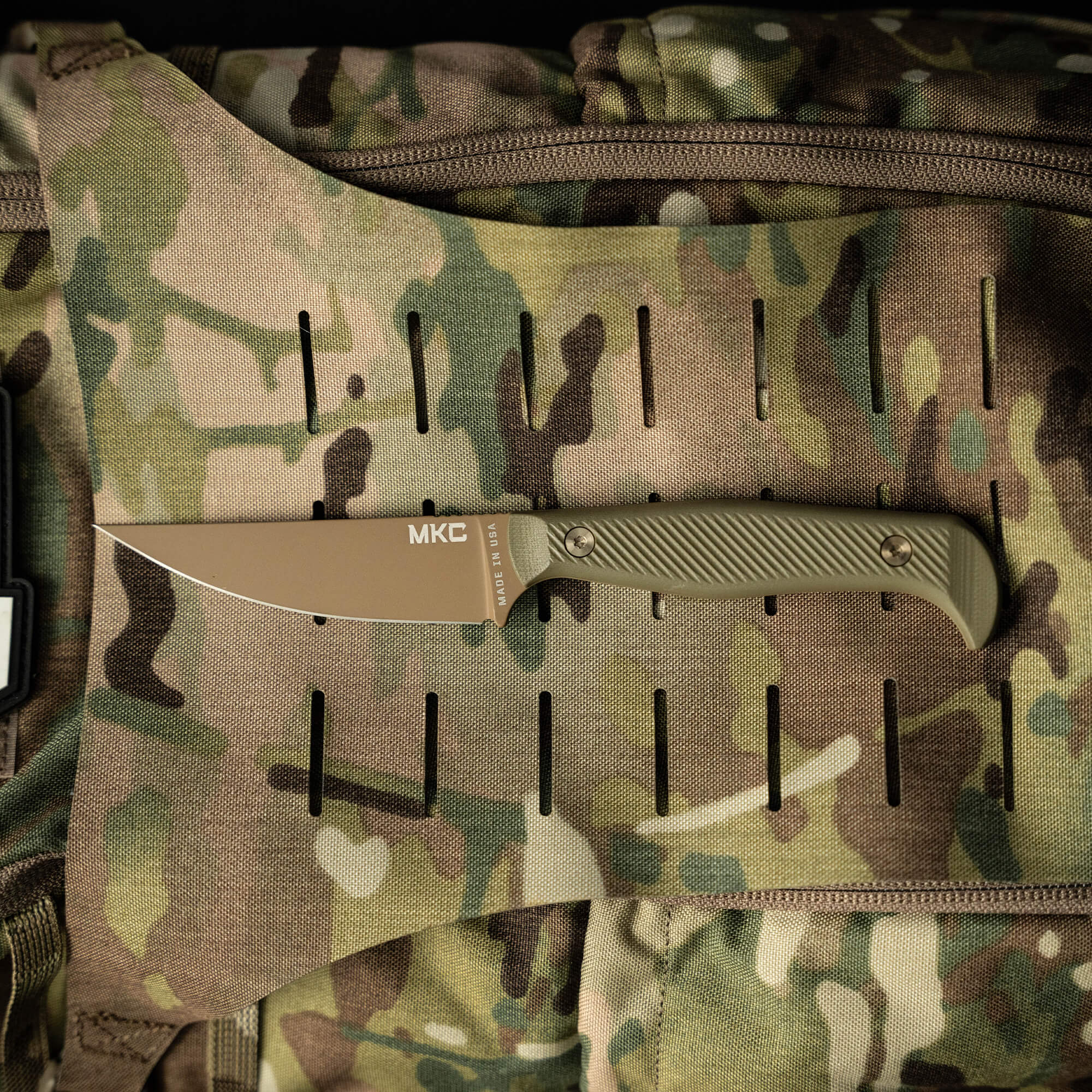 Alt text: "Battle Goat tactical knife in coyote olive drab color with a fixed blade, displayed on a camouflage fabric background featuring MOLLE webbing."
