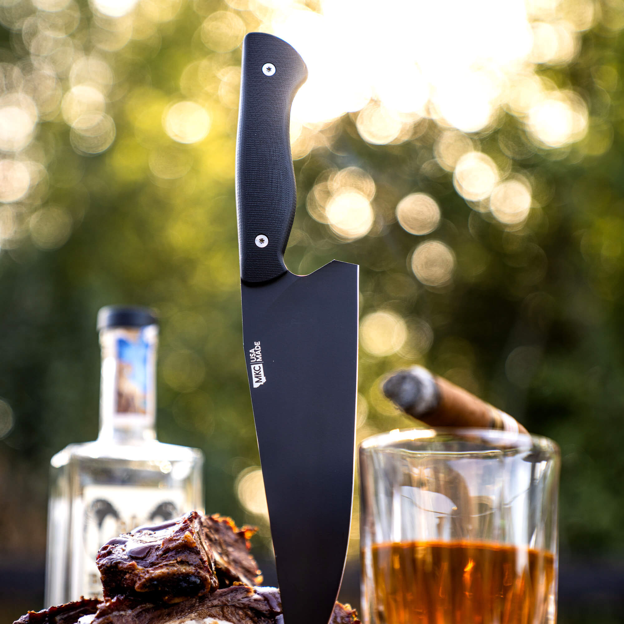Alt text: "Bighorn Chef black chef's knife vertically positioned against a blurred natural background, with its black handle and sharp matte blade prominently displayed. The knife is showcased alongside gourmet grilled meat, a glass of whiskey, and a lit cigar,
