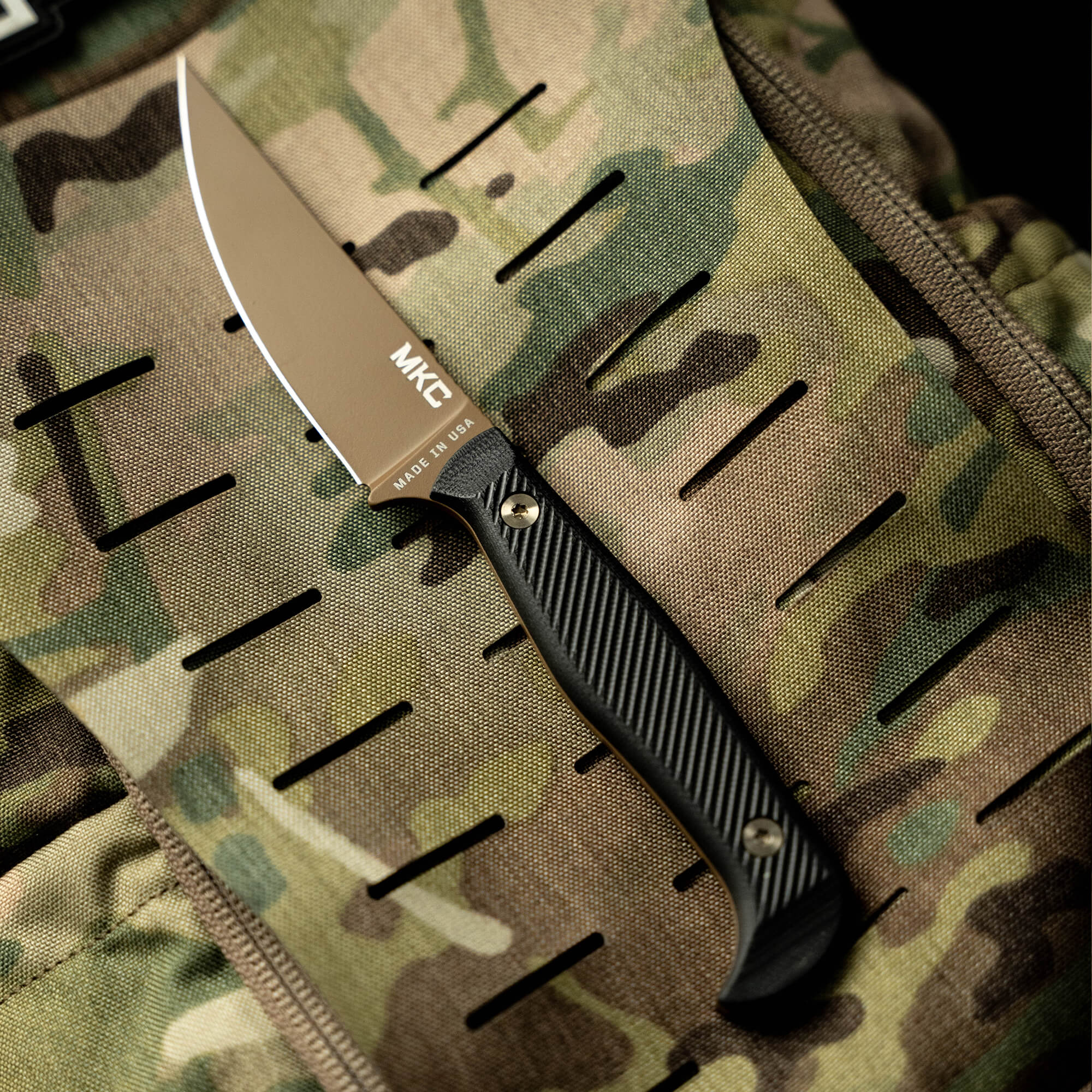 Alt text: Close-up of an MKT Battle Goat folding knife with a satin-finished blade and black handle, displayed against a coyote brown and black camouflage background.