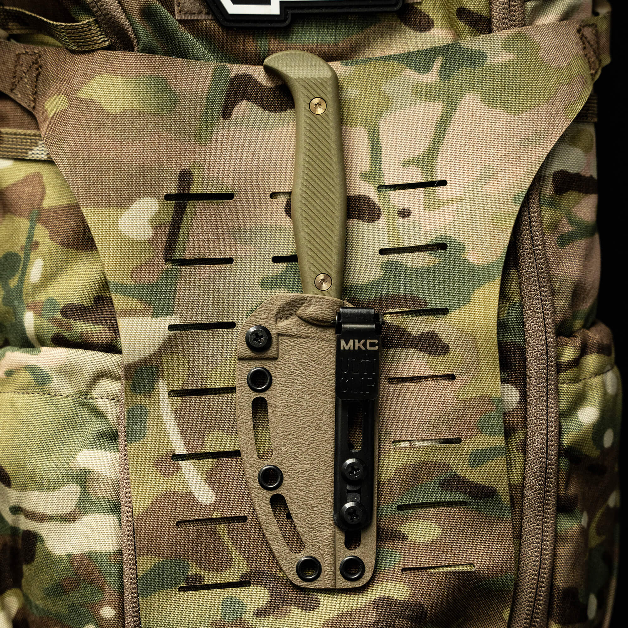 Alt text: Close-up view of a BATTLE GOAT tactical knife in coyote-olive drab color, secured on a camouflaged military backpack.