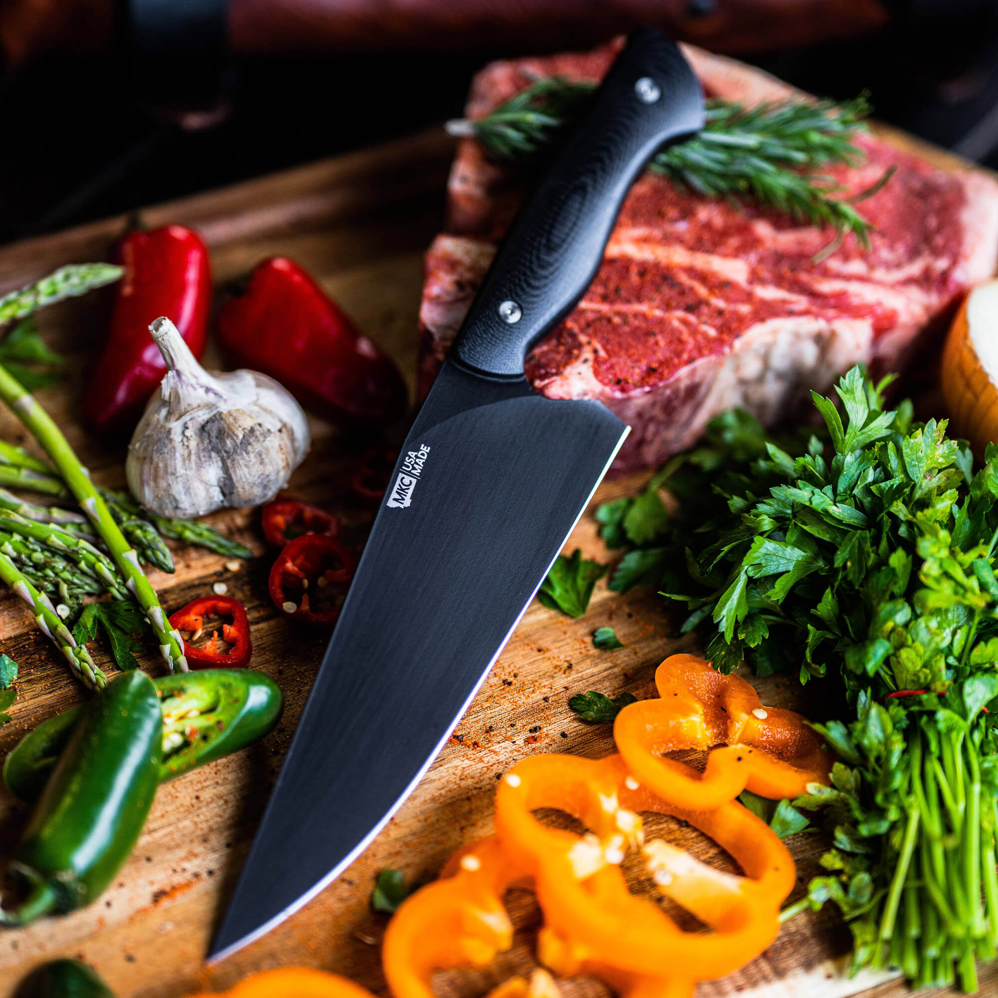 Alt text: "Bighorn Chef black chef's knife displayed on a wooden cutting board surrounded by fresh ingredients including red meat, various peppers, asparagus, and herbs."