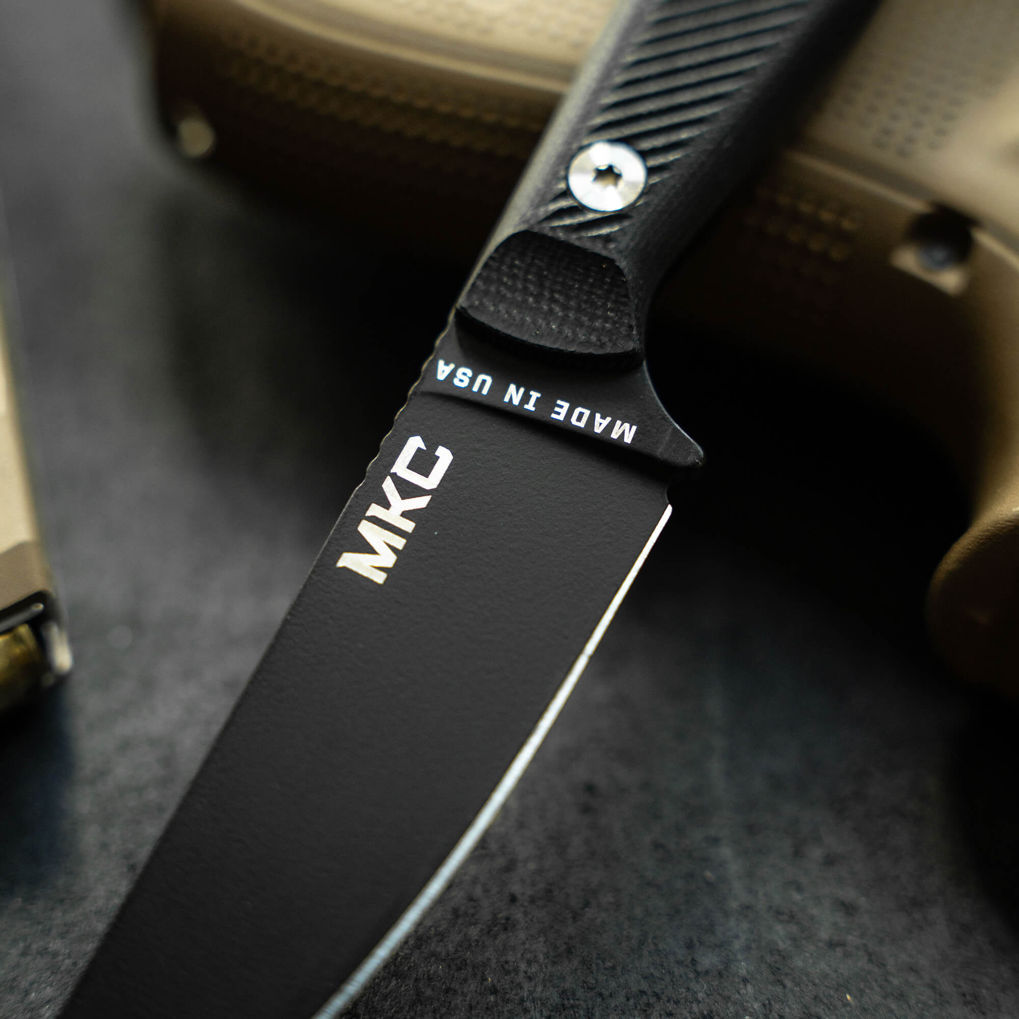Close-up of a black MKC Battle Goat knife with white text on the blade, set against a textured background.
