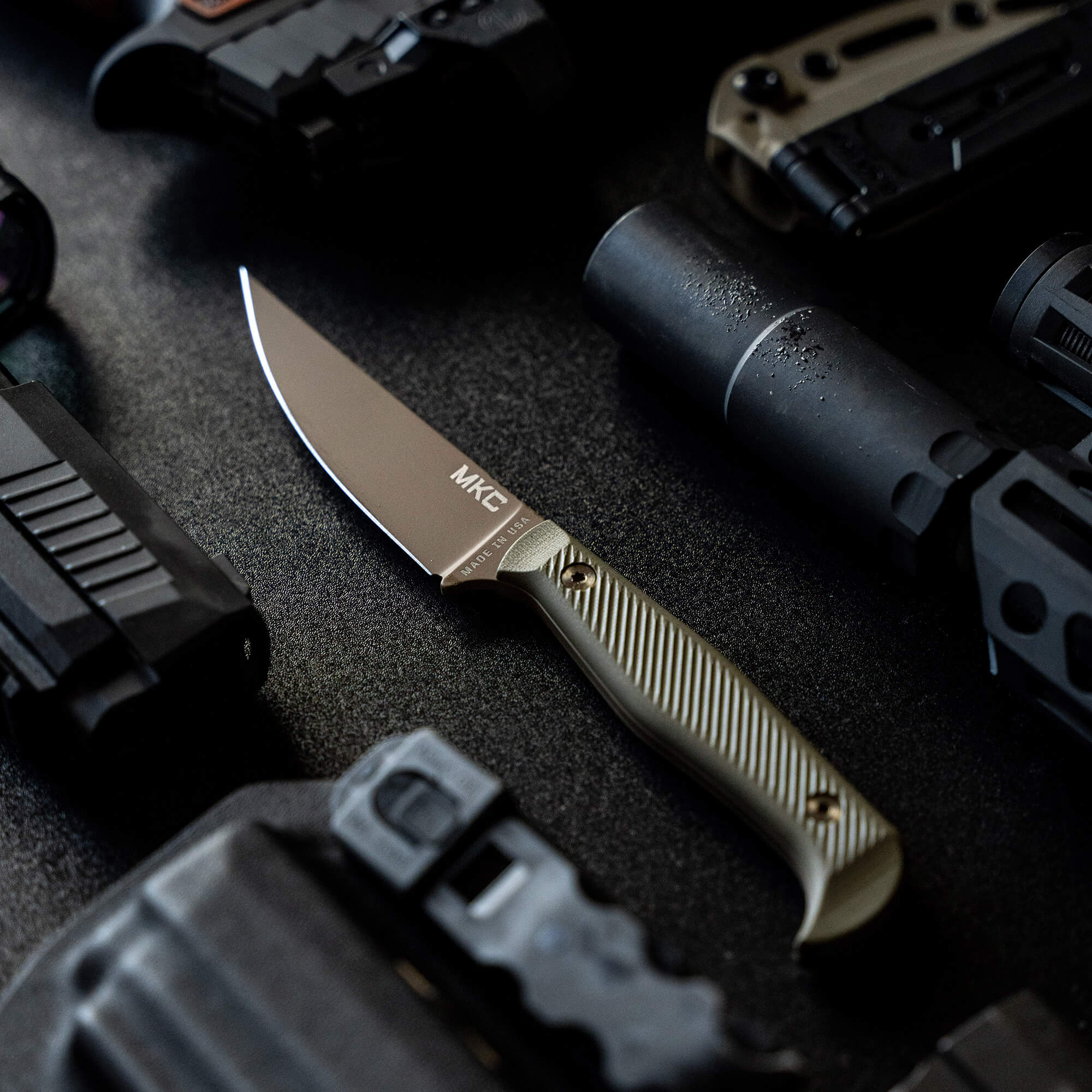 Alt text: "Close-up view of the BATTLE GOAT tactical knife in Coyote/Olive Drab, featuring a smooth tan handle and a sleek, straight-edged steel blade, surrounded by various tactical gear including optical sights and rugged equipment on
