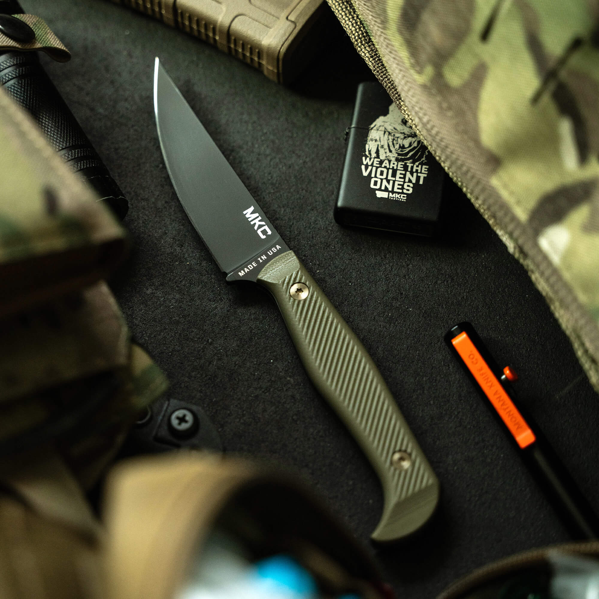 Alt Text: "MKC Battle Goat tactical knife with black blade and olive drab handle, displayed on a tactical gear background including a Zippo lighter and other outdoor equipment."