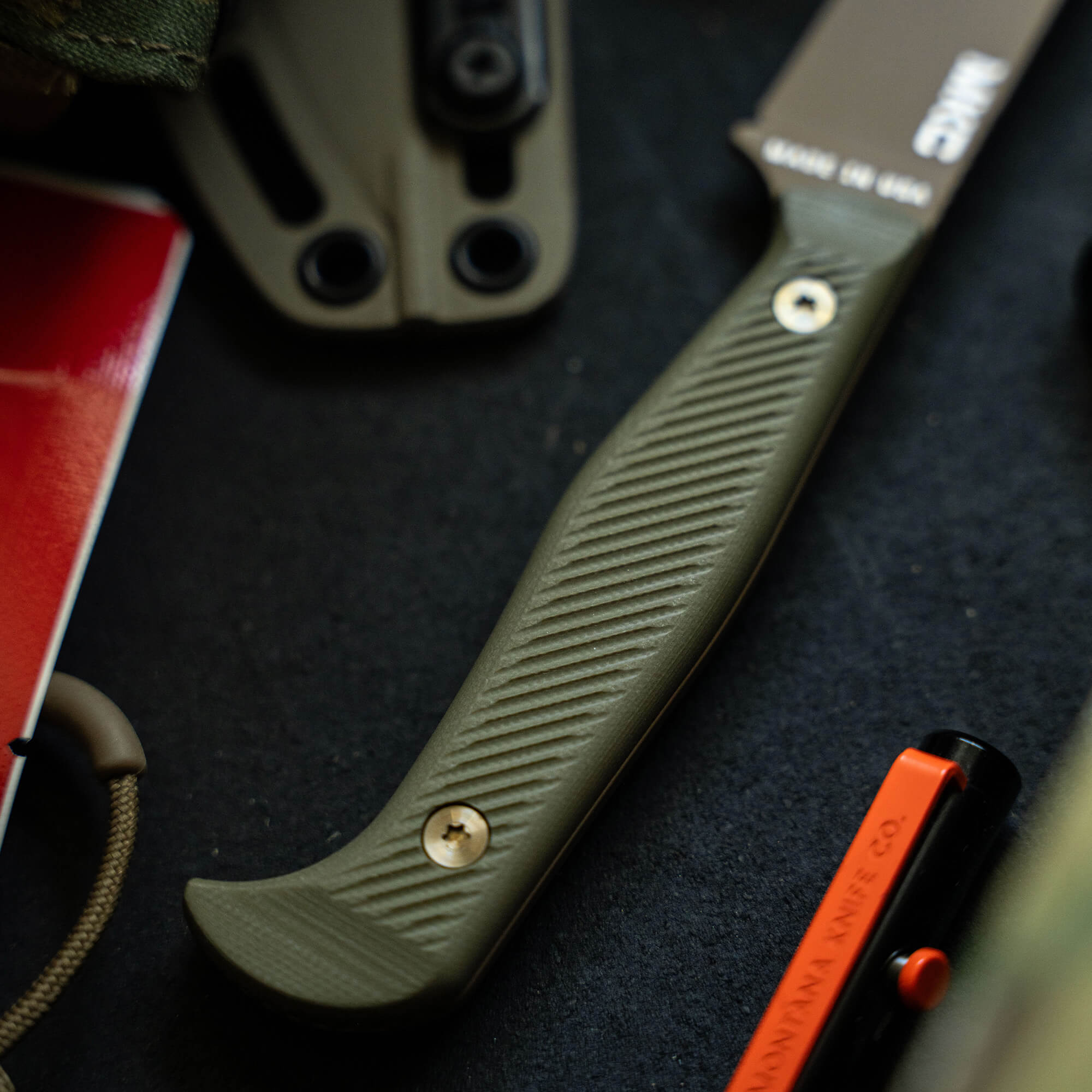 Close-up of a BATTLE GOAT fixed blade knife in Coyote/Olive Drab with a textured handle, showcased alongside tactical gear on a dark background.