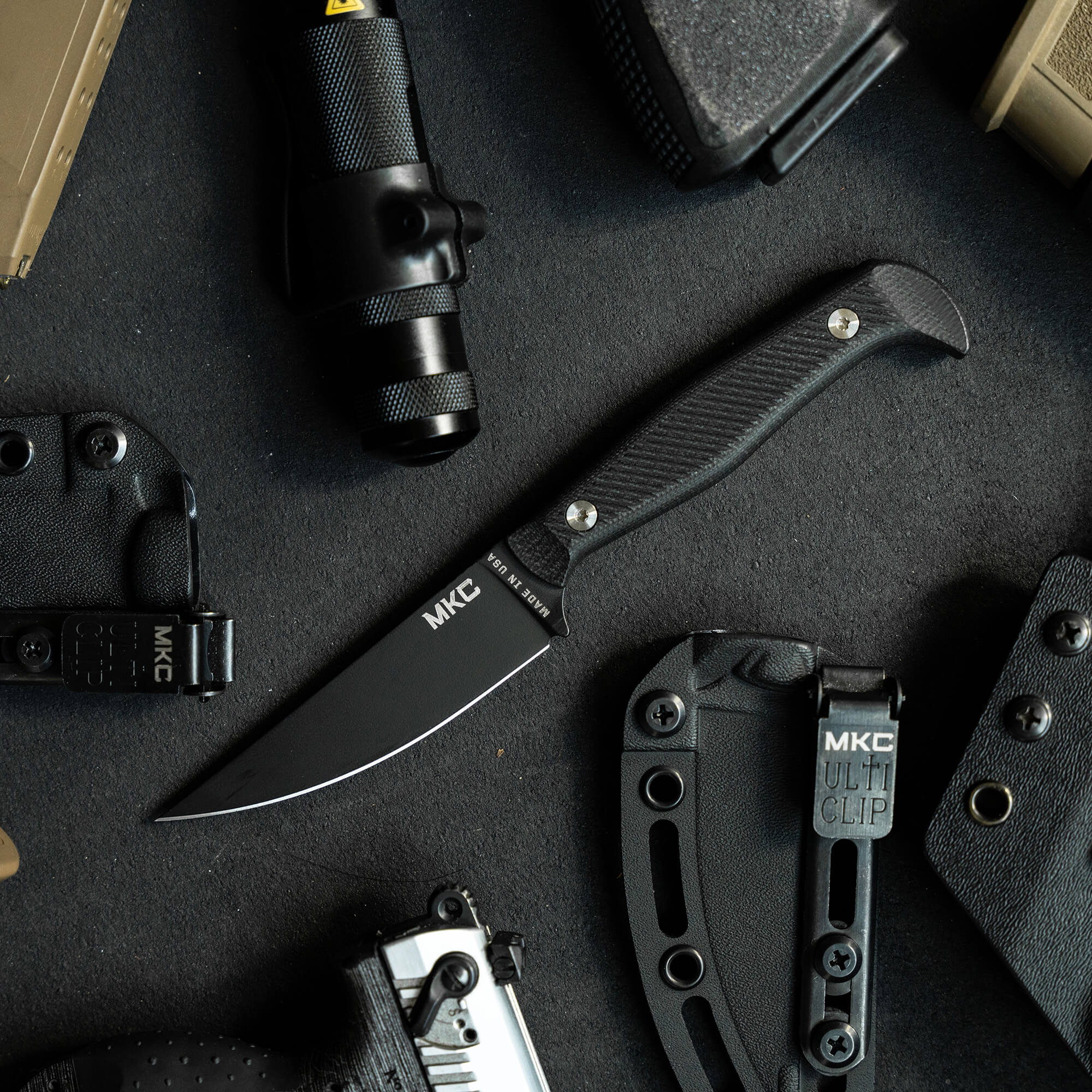 Alt text: "Black MKC Battle Goat tactical knife with a curved handle and sharp blade, displayed alongside tactical gear including a flashlight, holster, and multi-clip on a dark surface."