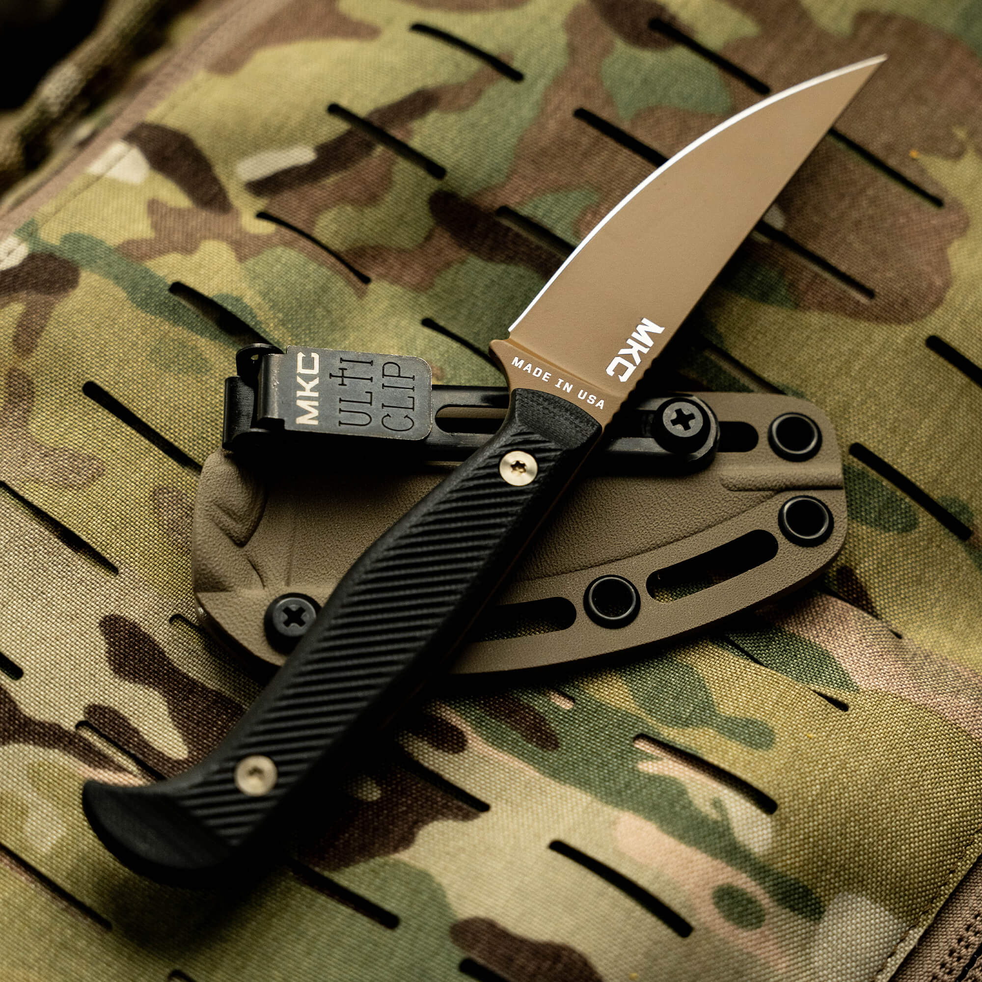 Alt text: Close-up of a Battle Goat tactical knife in coyote black color with a serrated blade, displayed on a camouflaged fabric background. The knife features a prominent MKC logo on the belt clip and is labeled "Made in USA