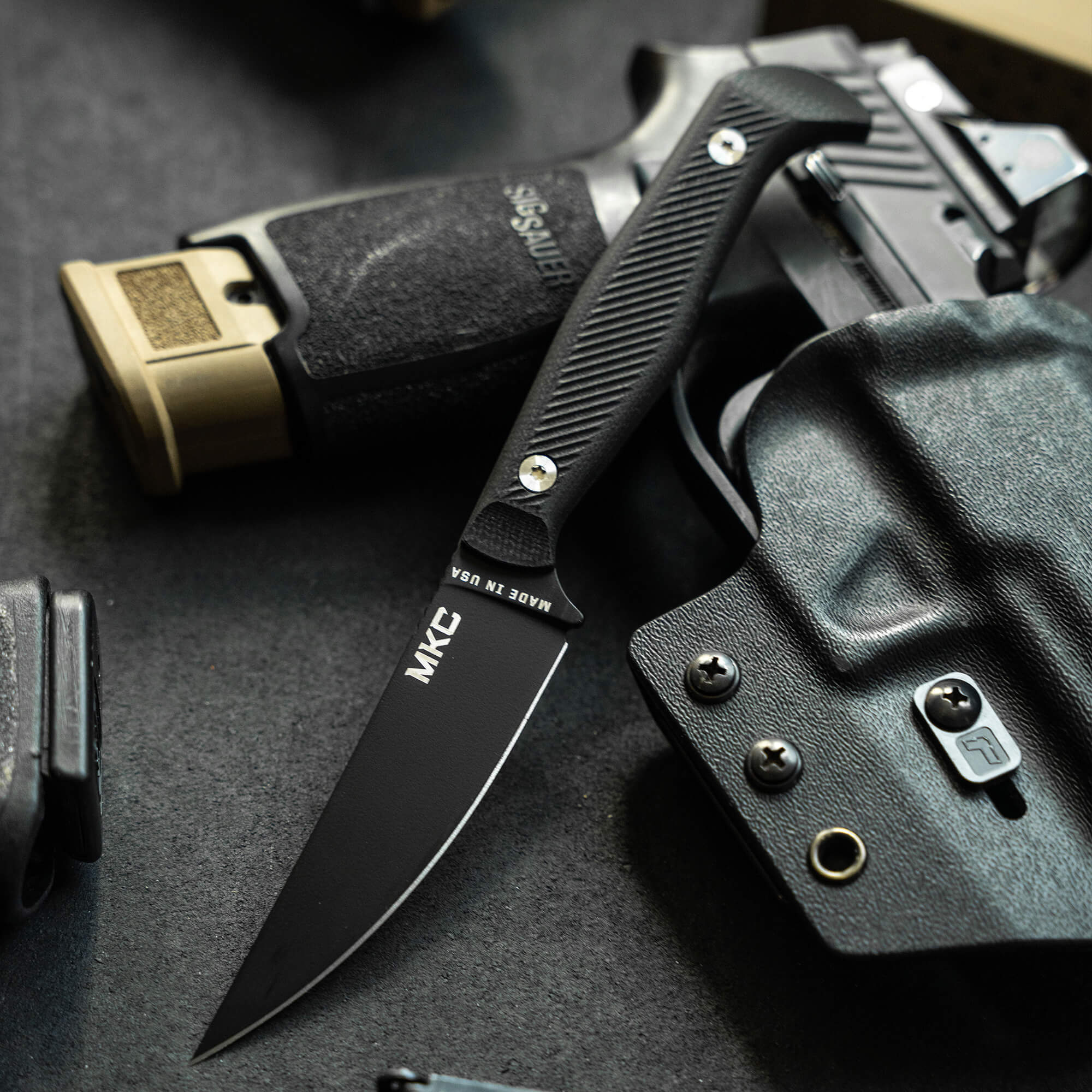 Alt text: Close-up of a black MKC tactical knife with a pointed tip and textured handle alongside a partially visible SIG SAUER firearm and a black holster on a dark surface.