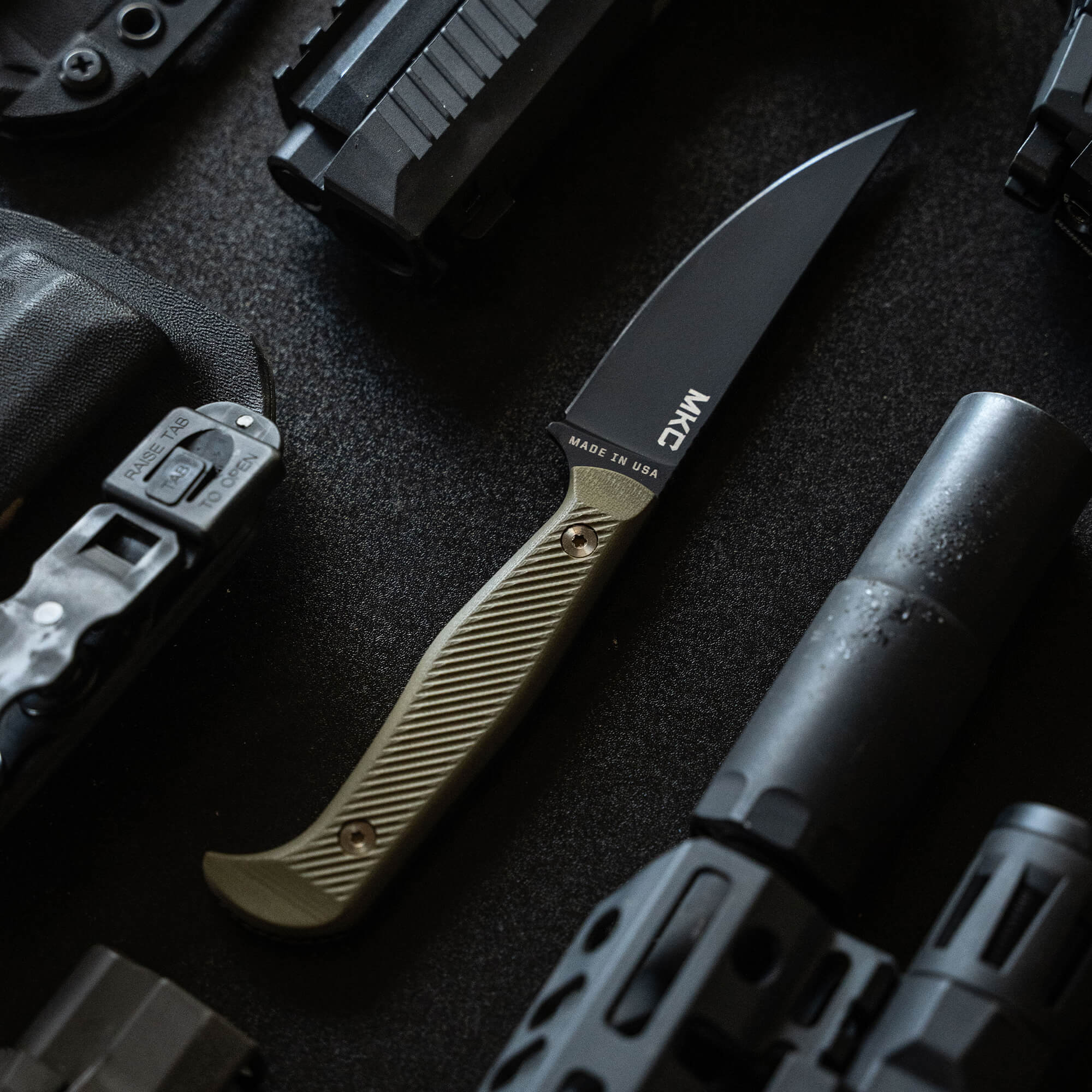 Alt text: Close-up view of a MKM Battle Goat tactical knife with a black blade and olive drab handle, displayed among tactical gear including firearm magazines and parts, showcasing its rugged design and utility.