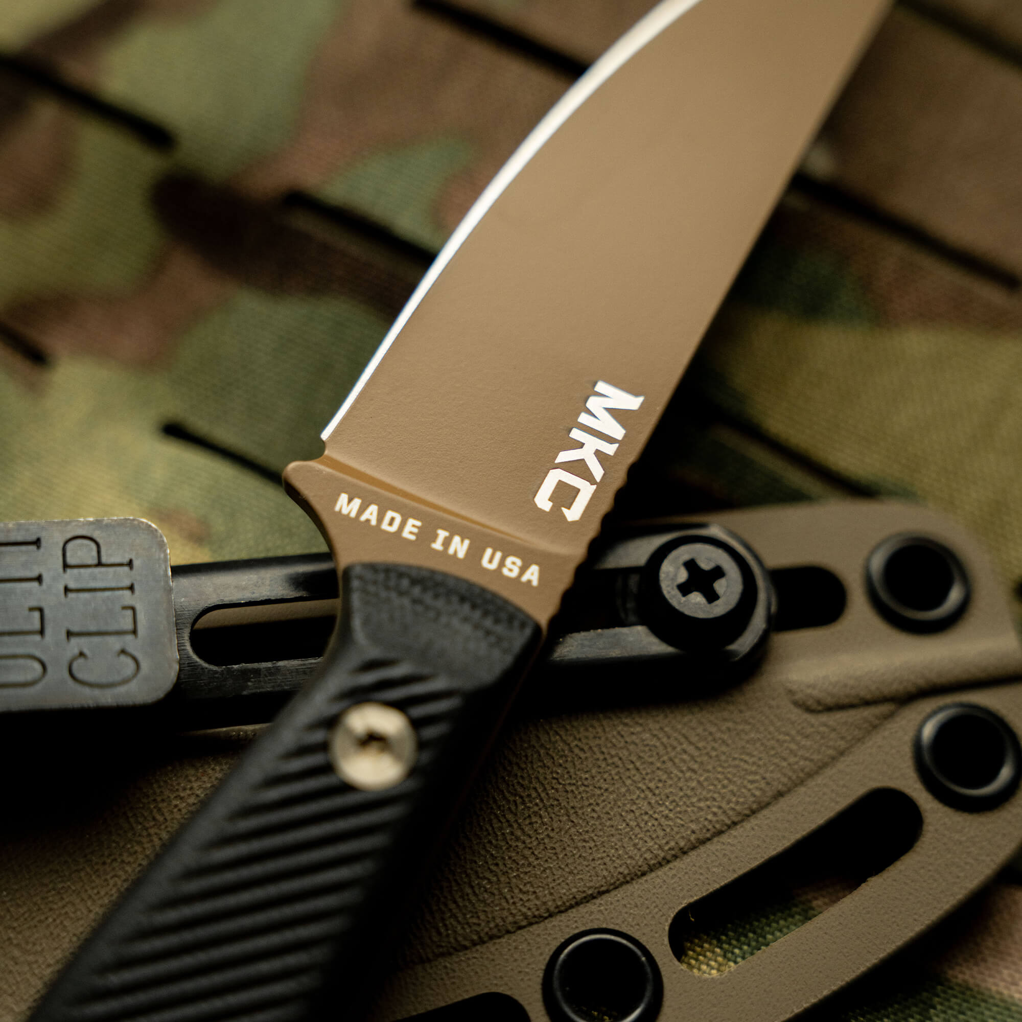 Alt text: Close-up of a MKC Battle Goat fixed-blade knife in coyote black, featuring a textured handle, sharp blade, and "Made in USA" mark, positioned against a camouflaged background.