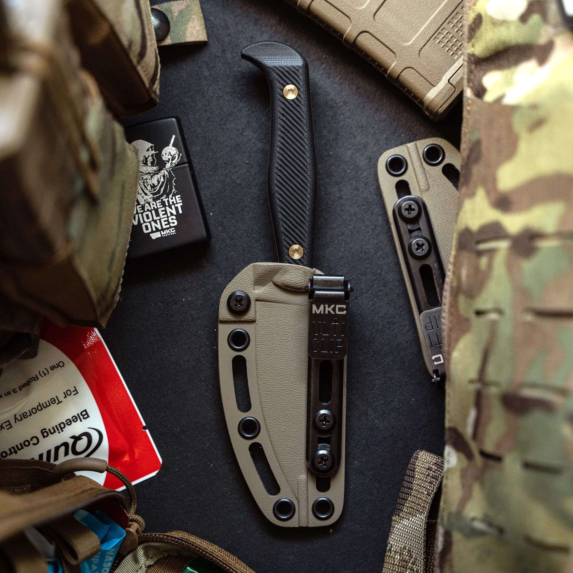 Alt text: A tactical fixed-blade knife with a black handle and coyote tan sheath, displayed amidst military gear including a camouflage pattern backpack, zippo lighter, and other outdoor equipment.