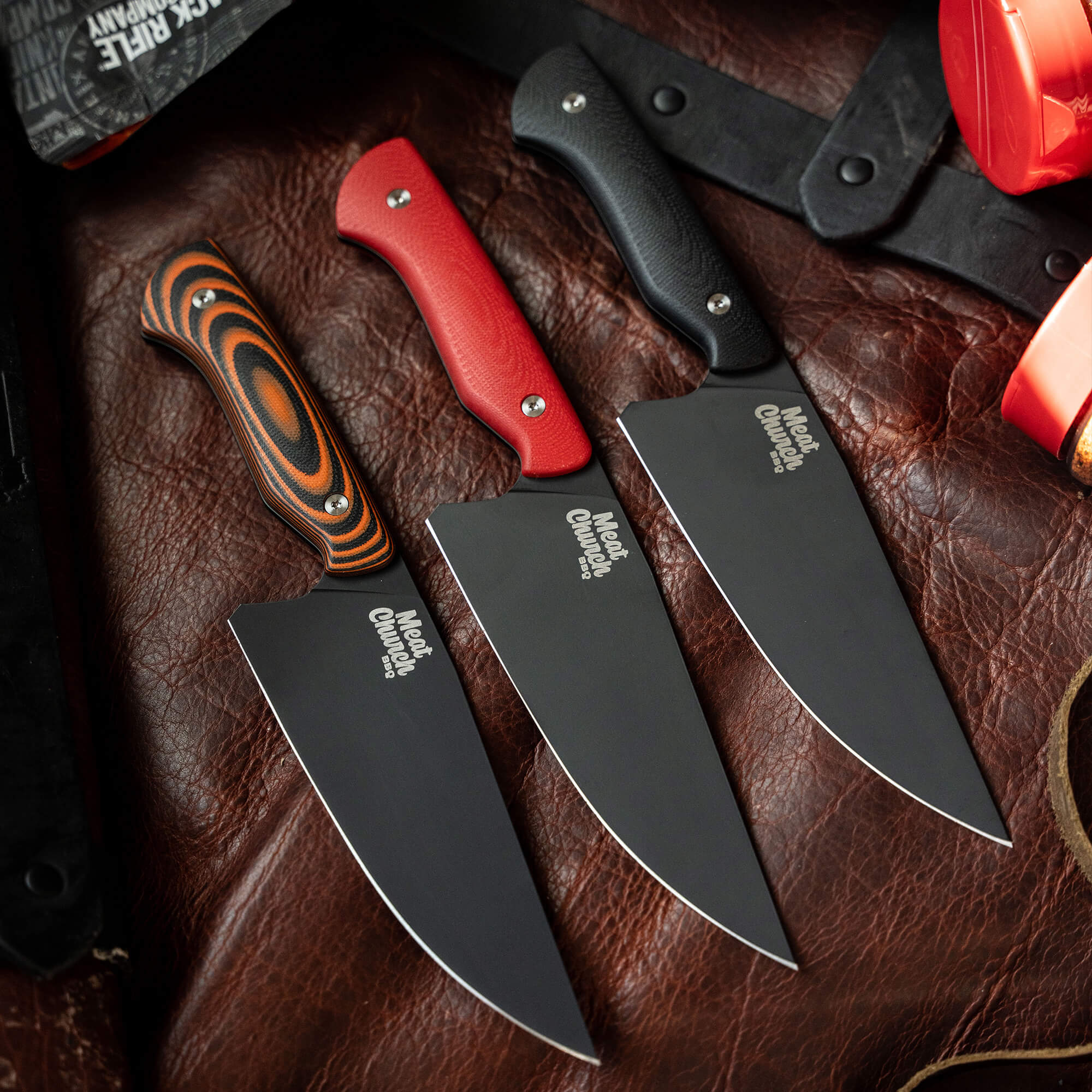 Alt text: "An array of Meat Church chef knives on a vintage leather background, featuring black blades with Meat Church logo and handles in orange swirl, red, and black colors. Each knife has a thick, sturdy blade, ideal for culinary precision."