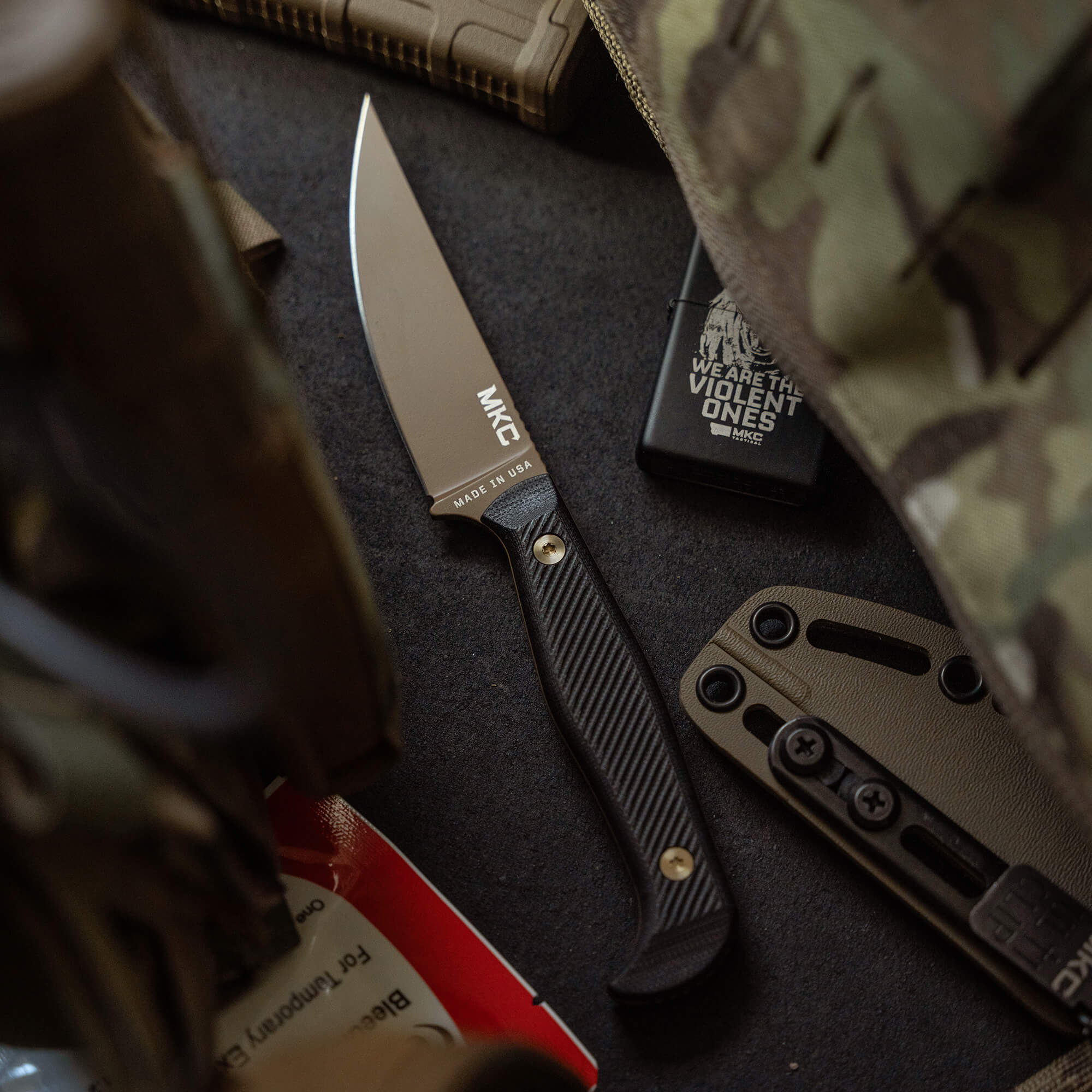 Alt text: "Close-up of a BATTLE GOAT fixed blade knife in Coyote Black color with a textured handle, displayed alongside tactical gear including a camouflage bag, survival tools, and branded merchandise on a dark surface."